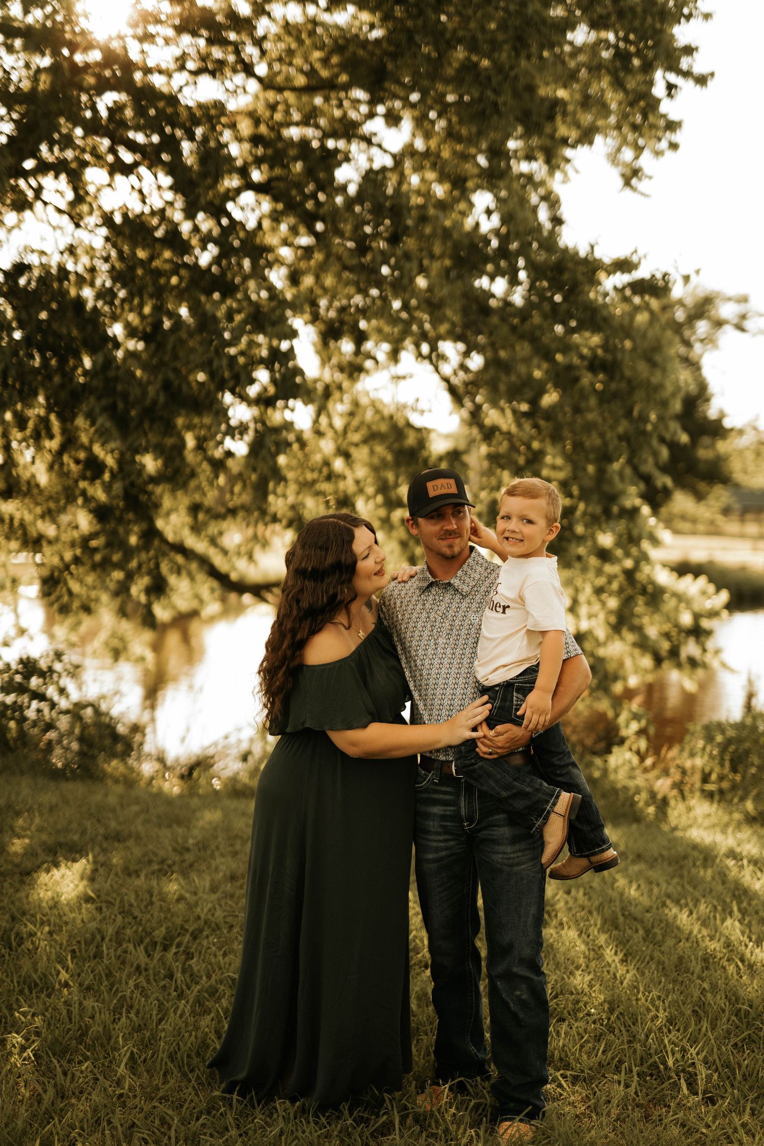 Family of three poses together outdoors with golden sunlight filtering through trees behind them.