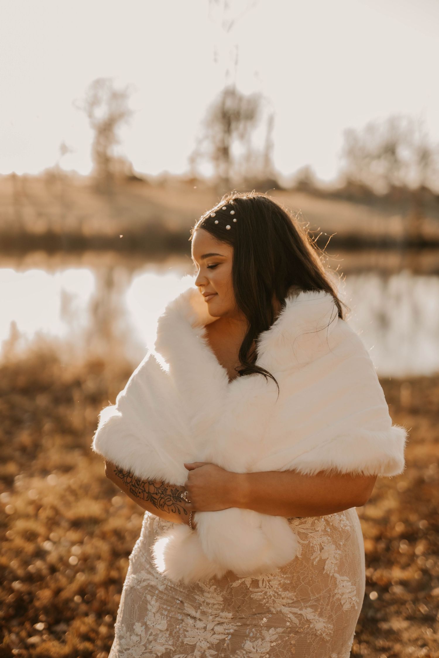 A bride in a white fur wrap smiles during golden hour by a lake.