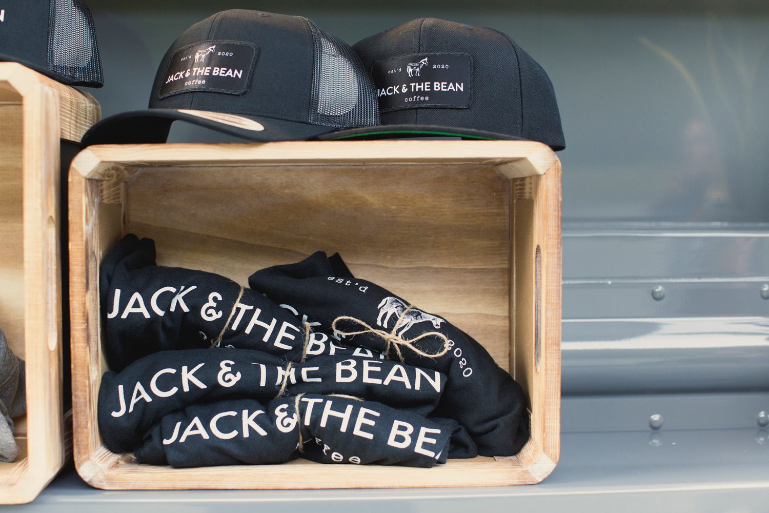 Black baseball caps and t-shirts with Jack & The Bean logo merchandising displayed in wooden box.