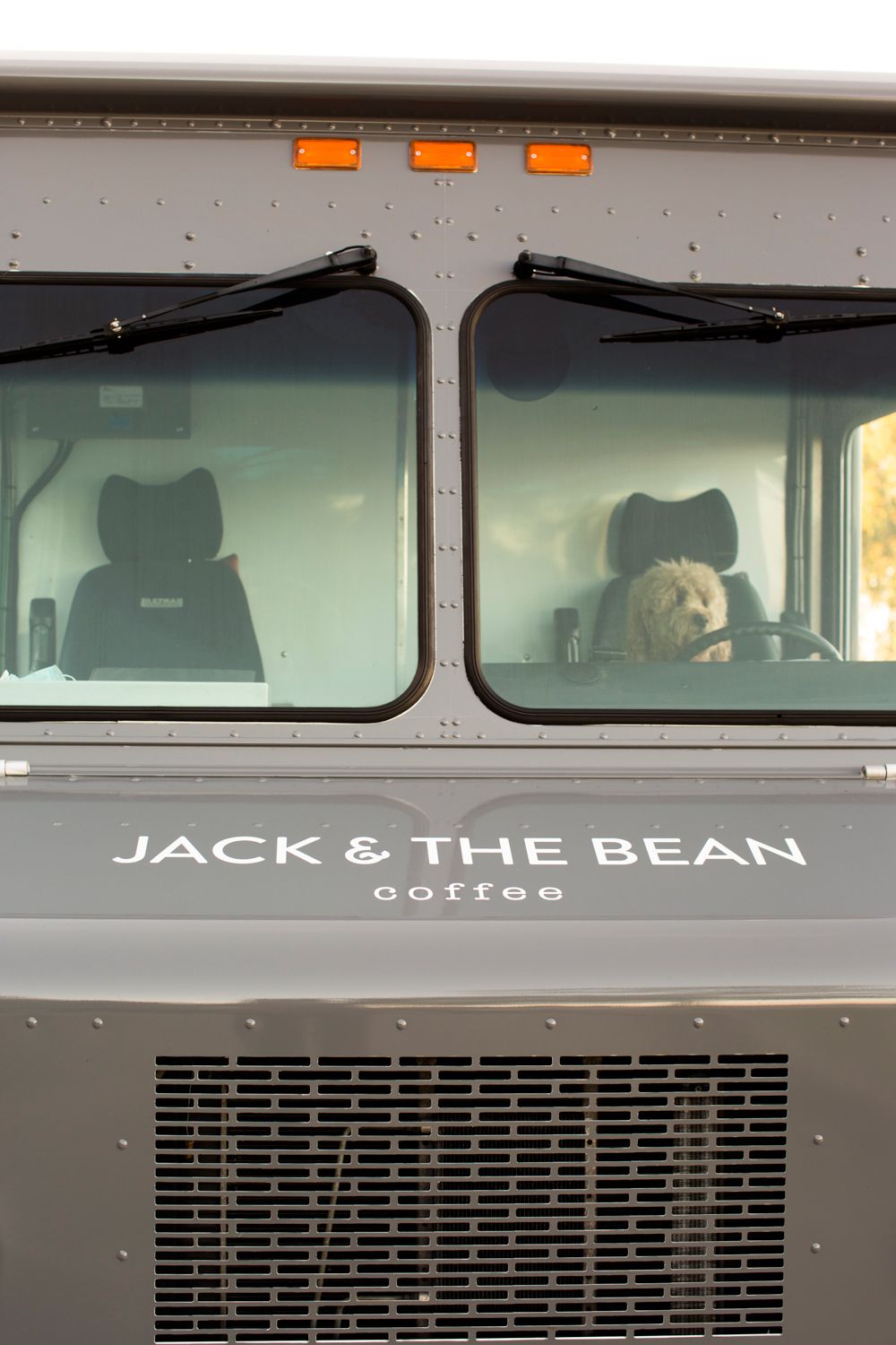 Front windshield view of a dog sitting in the driver's seat of a coffee delivery truck.