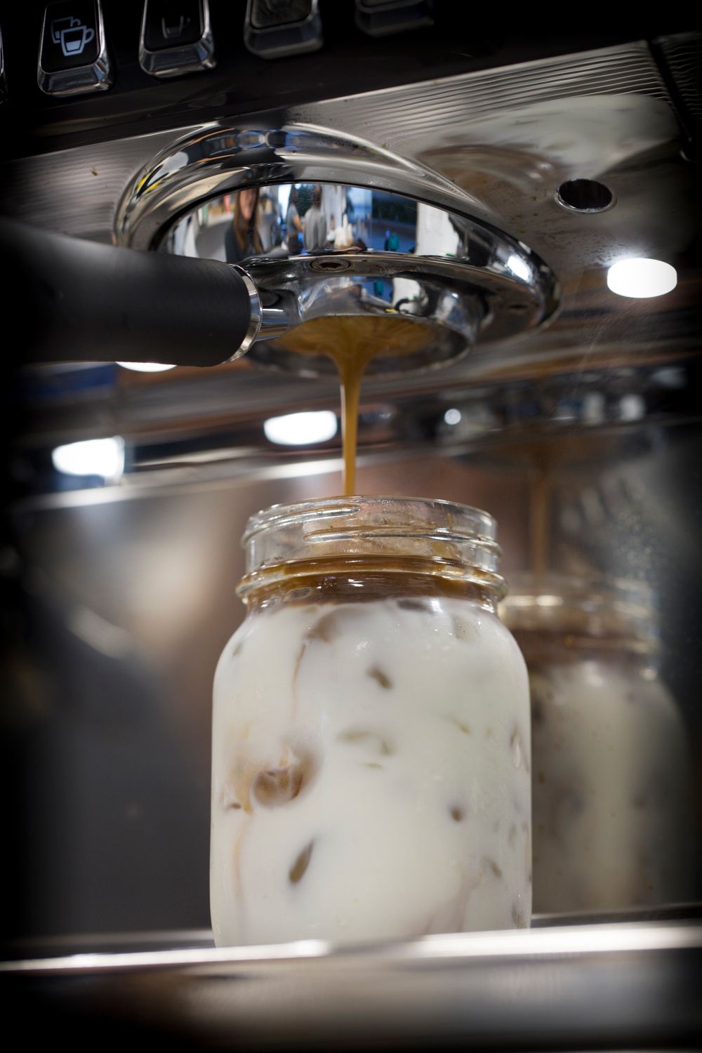 Coffee being poured into mason jar filled with cream under chrome espresso machine.