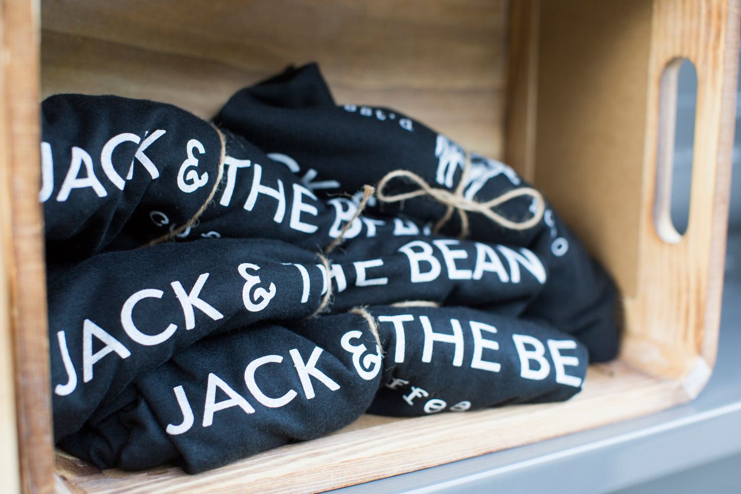 Folded black t-shirts with Jack & The Bean branding displayed in wooden crate.