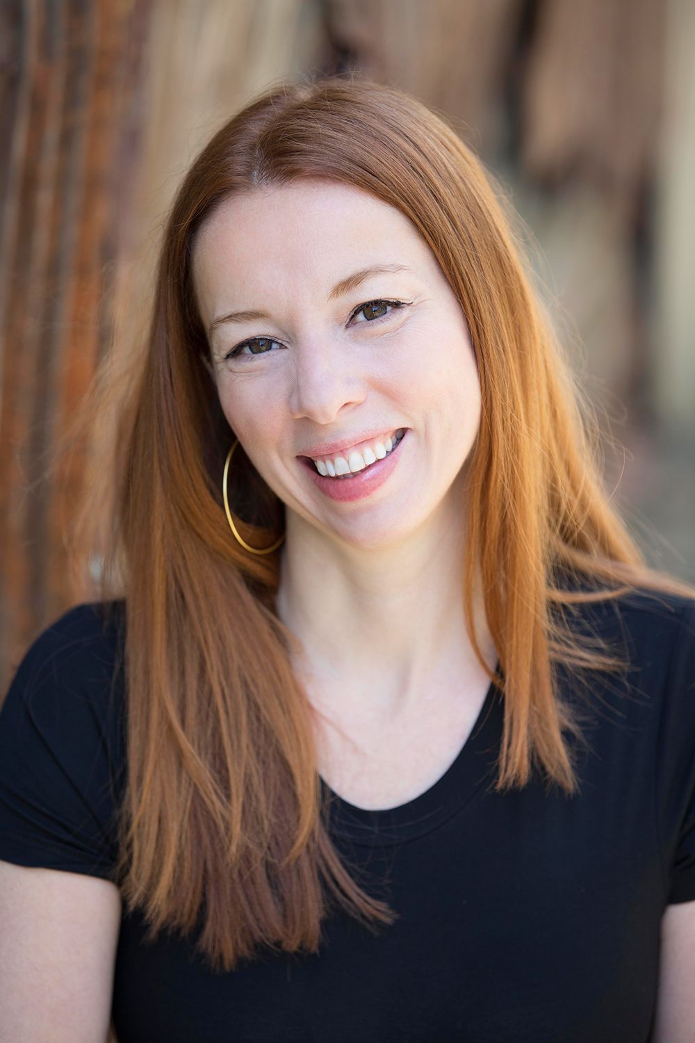Smiling woman with long red hair wearing gold hoop earrings and black top in outdoor setting.
