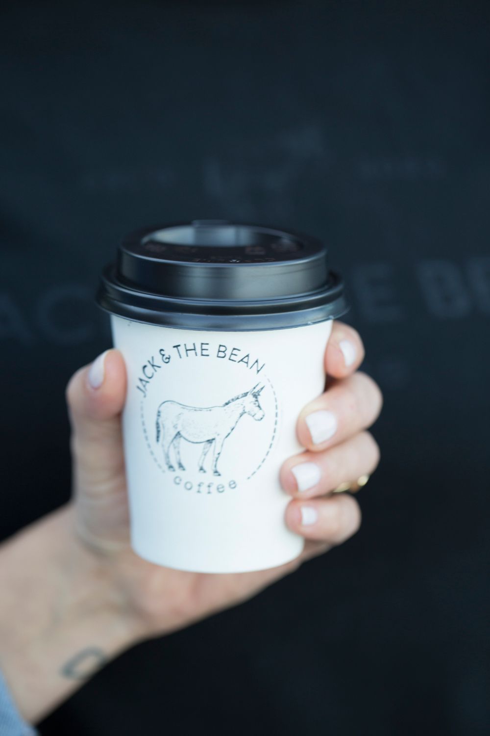 Close-up of hand holding white coffee cup with donkey logo against dark background.