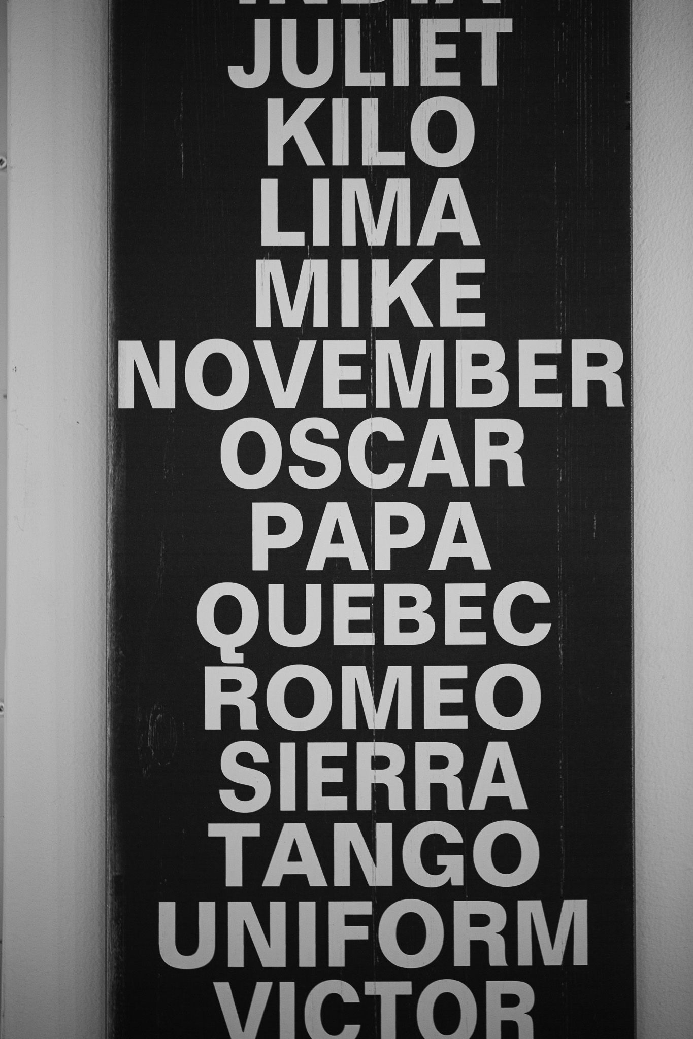 Black and white sign displaying NATO phonetic alphabet letters in vertical layout.