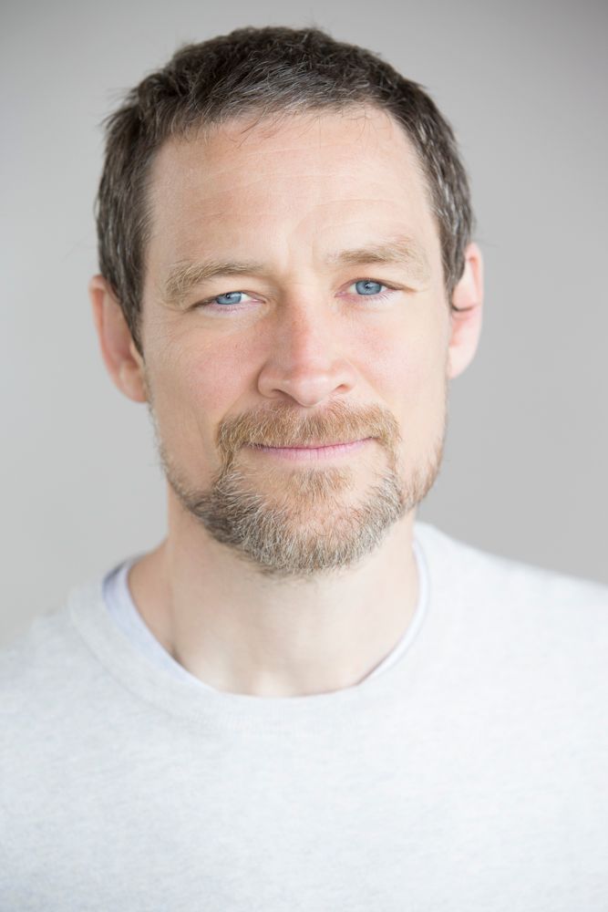 Portrait of a man with blue eyes and trimmed beard wearing a white t-shirt against a light background.