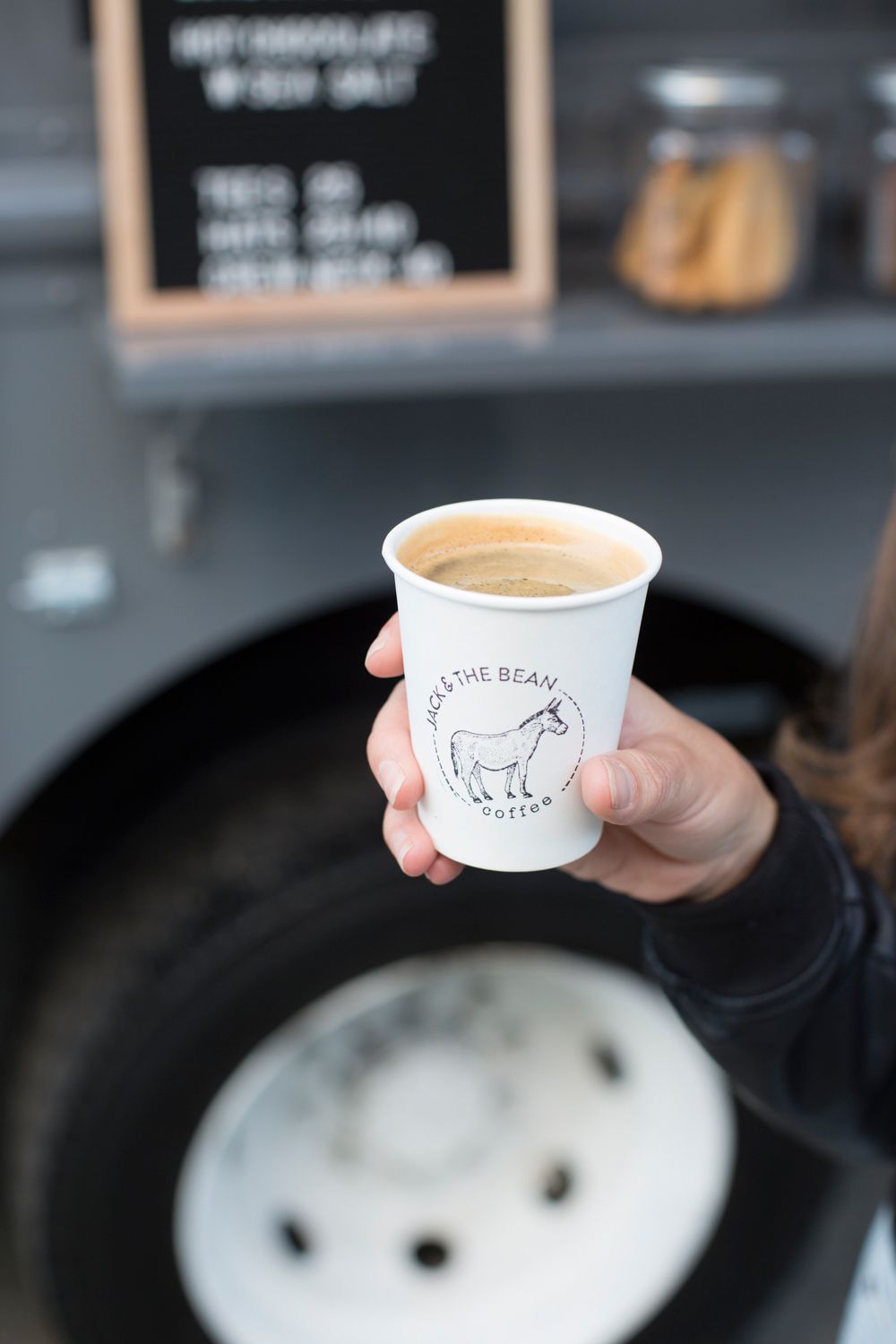Hand holding a white coffee cup with Jack & The Bean donkey logo against dark background.