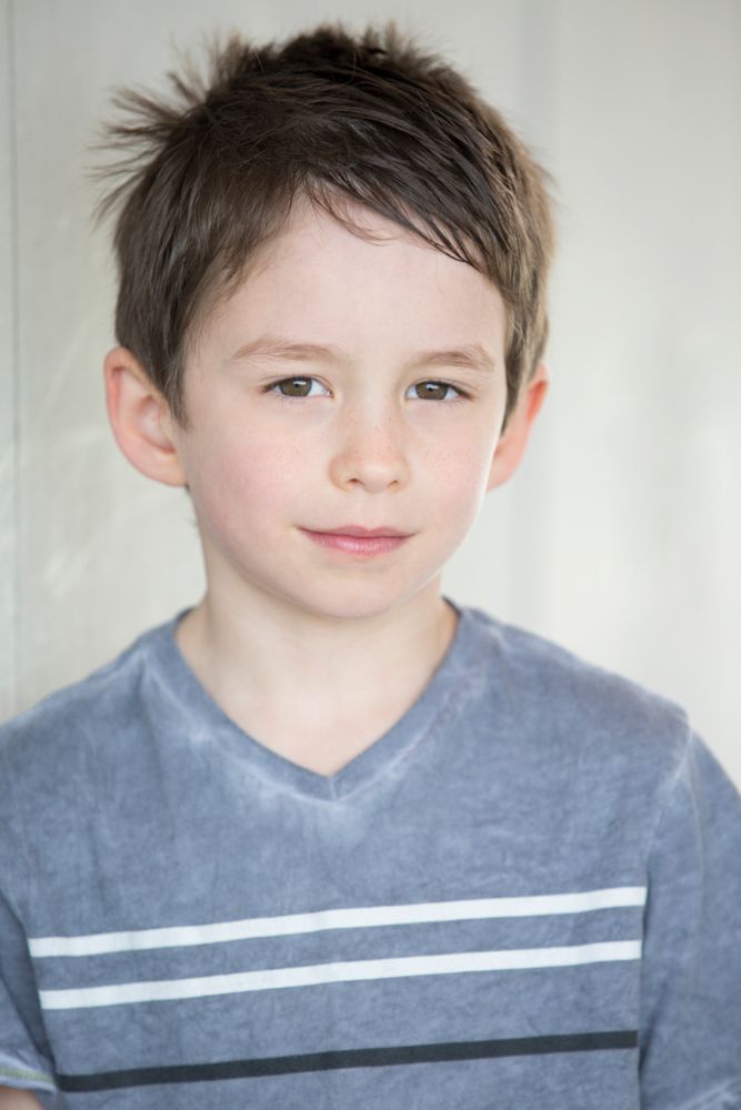 Young child in grey striped t-shirt smiling at camera against light background.