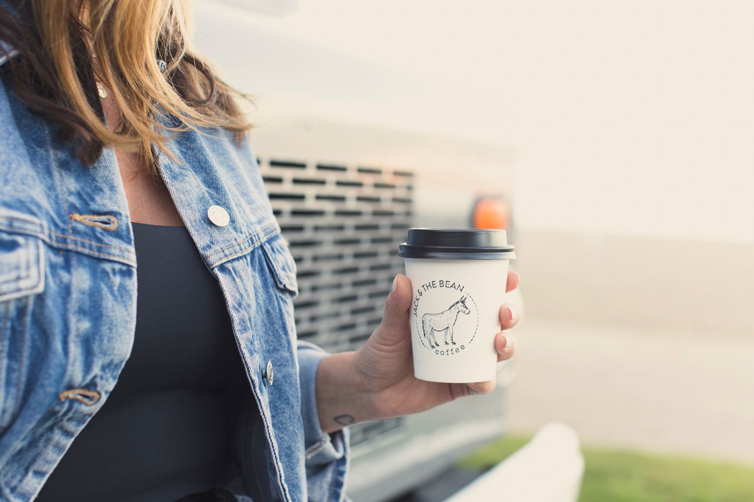 Coffee cup held by someone wearing denim jacket against light background.