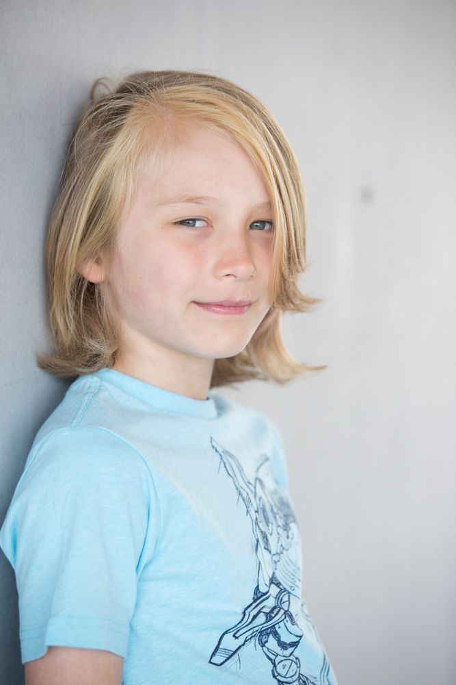 Young person with blonde hair wearing a light blue t-shirt with graphic design against a white wall.