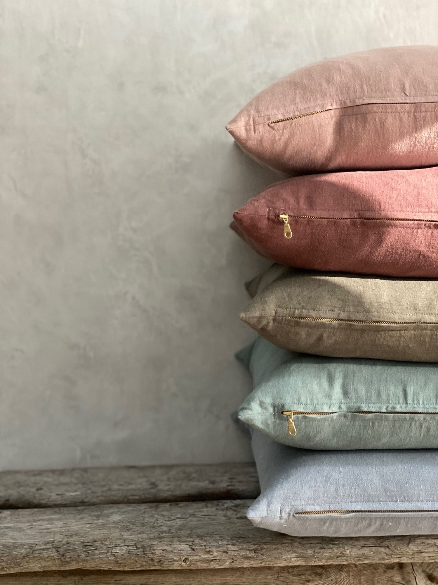 Stack of linen pillows in muted pink, beige and blue tones arranged on wooden surface.