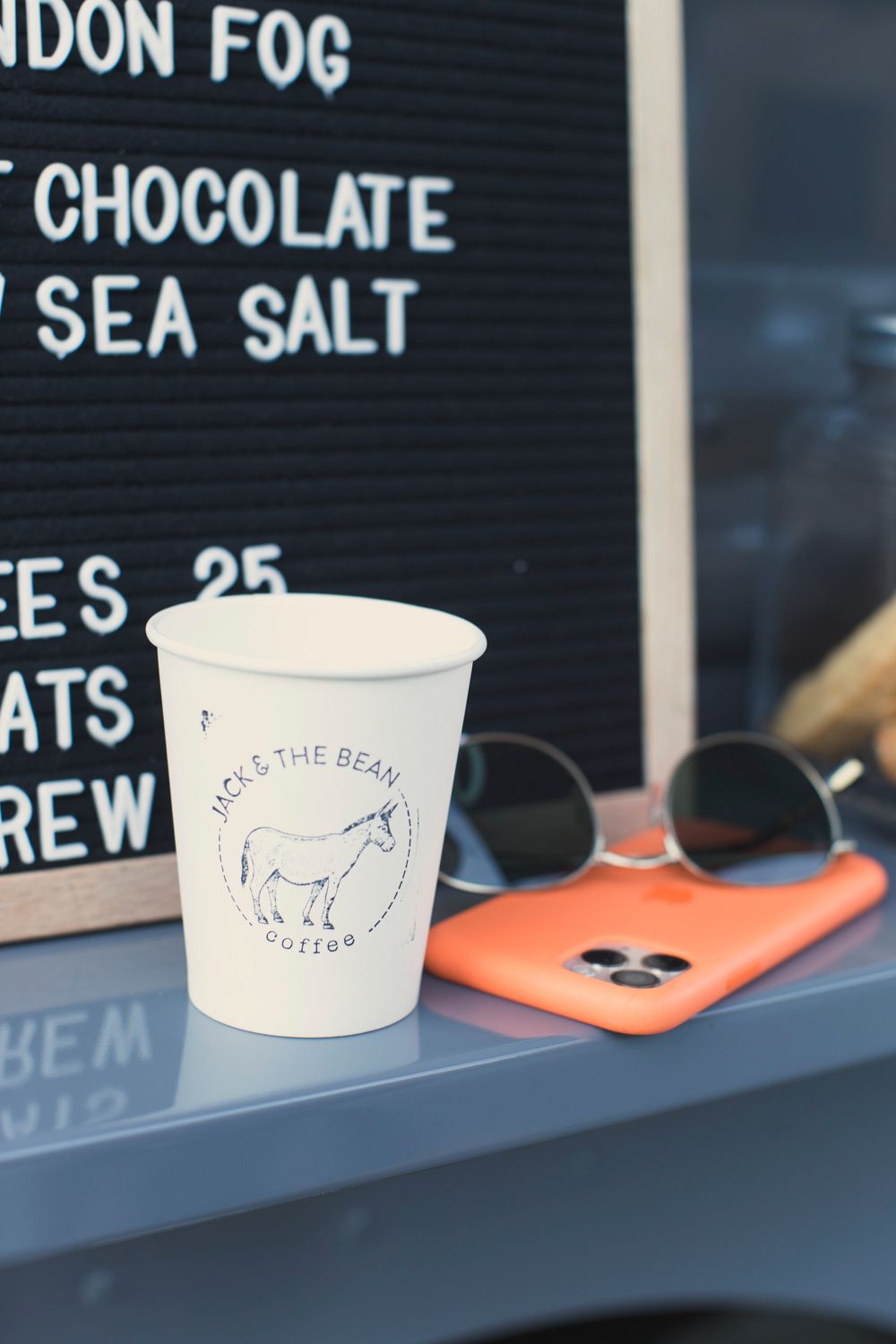 White paper coffee cup with Jack & The Bean logo sitting on counter next to menu board.