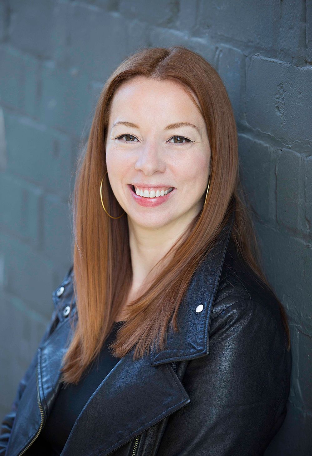 Portrait of a woman with long auburn hair wearing a black leather jacket against a dark wall.