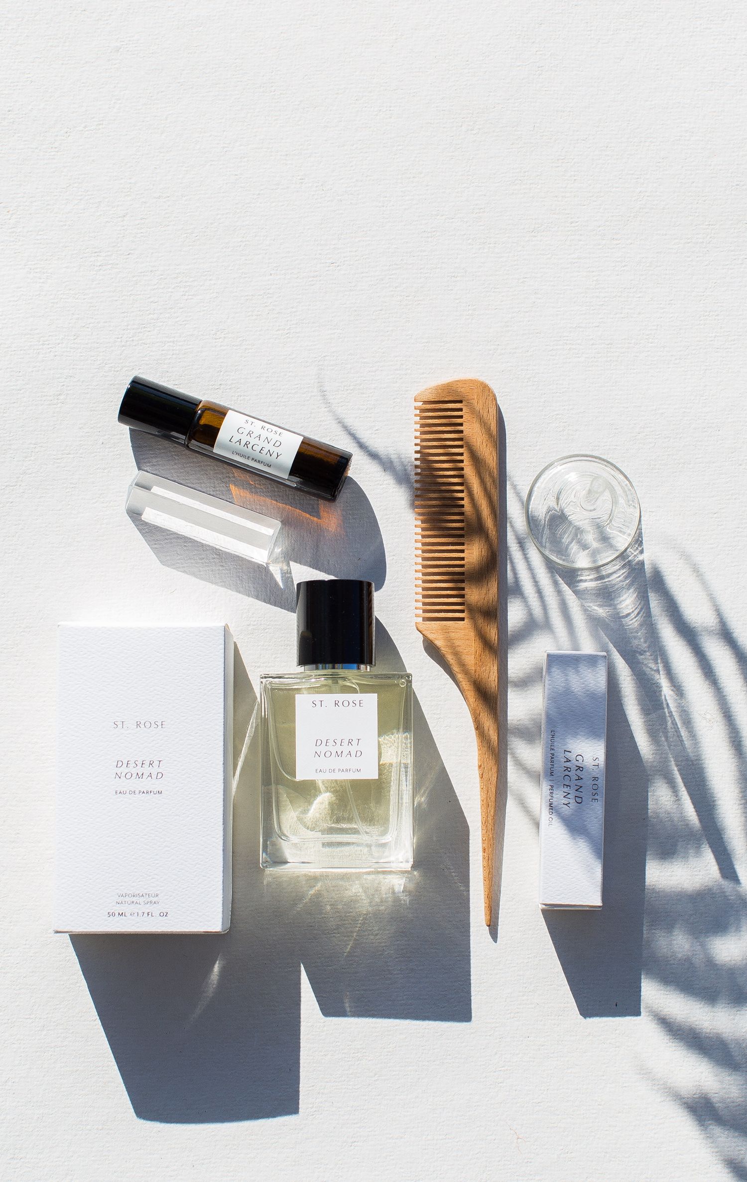 Perfume bottles and wooden comb arranged with palm leaf shadows on white surface.