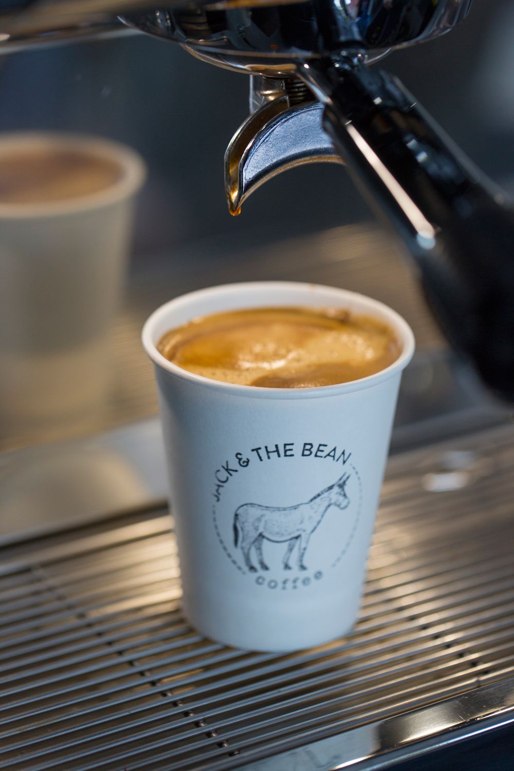Fresh espresso in a white paper cup with donkey logo sitting on metal coffee machine grating.