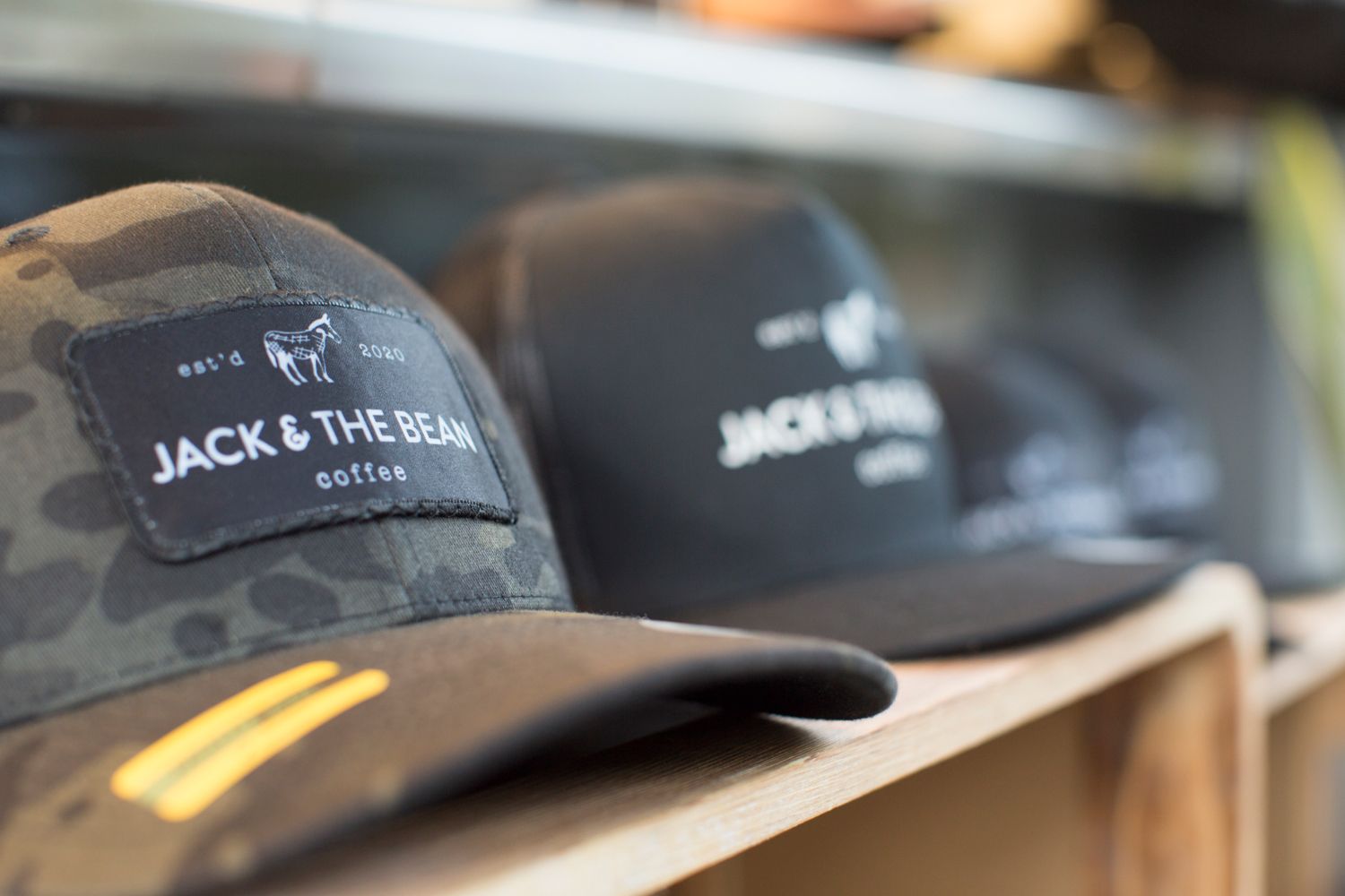 Camouflage and black baseball caps with Jack & The Bean coffee branding on retail display.