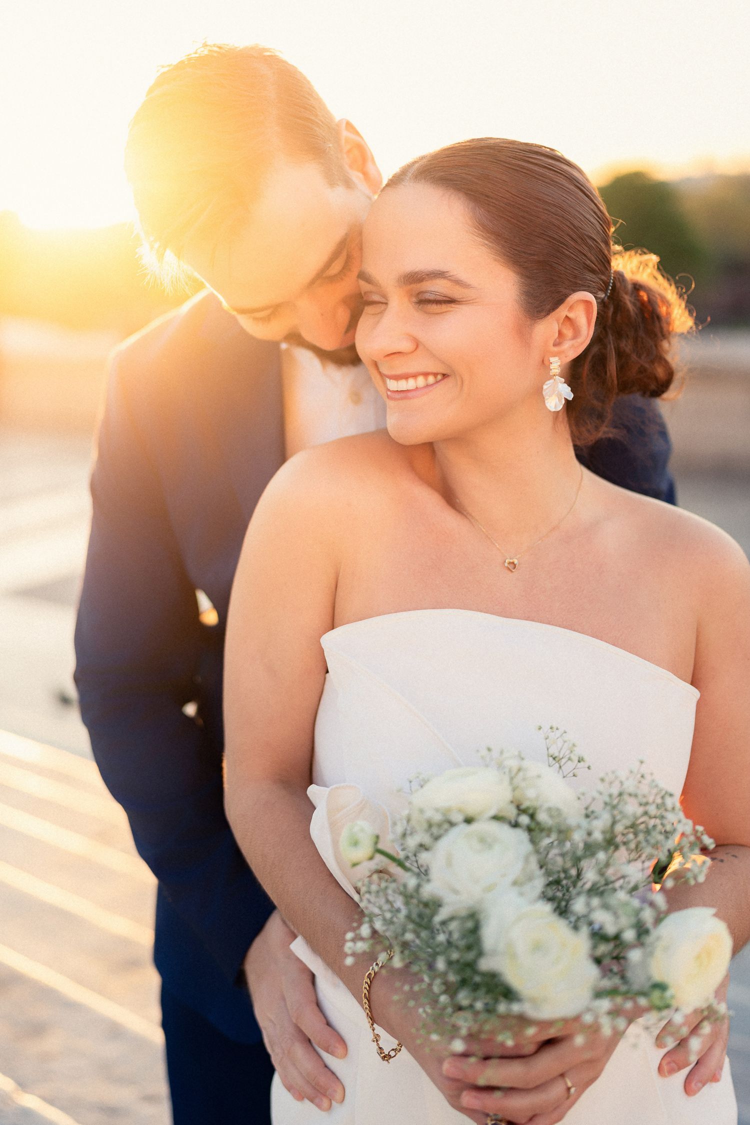 Romantic sunset wedding portrait featuring a couple embracing with white bouquet on beach.
