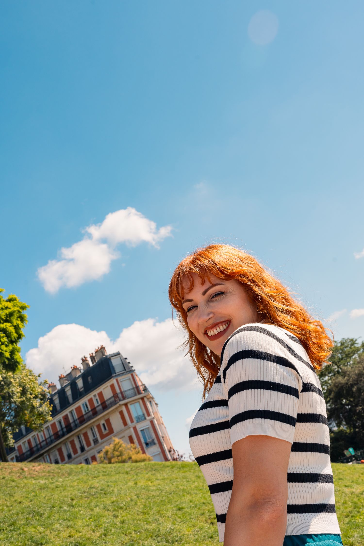 Someone in a striped shirt smiling against a blue sky with a French building in the background.