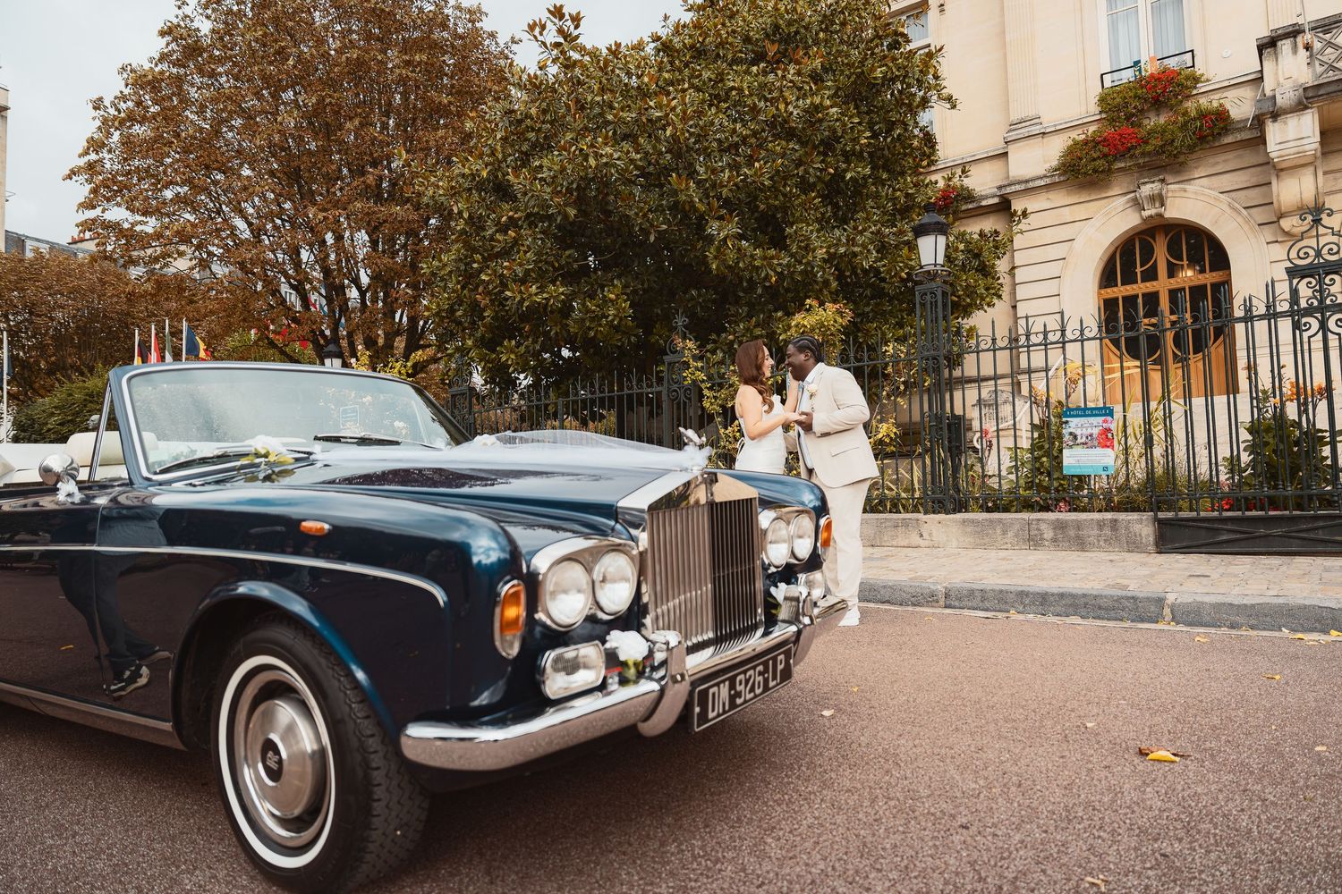 Classic blue vintage car parked in front of elegant stone buildings on a tree-lined street.