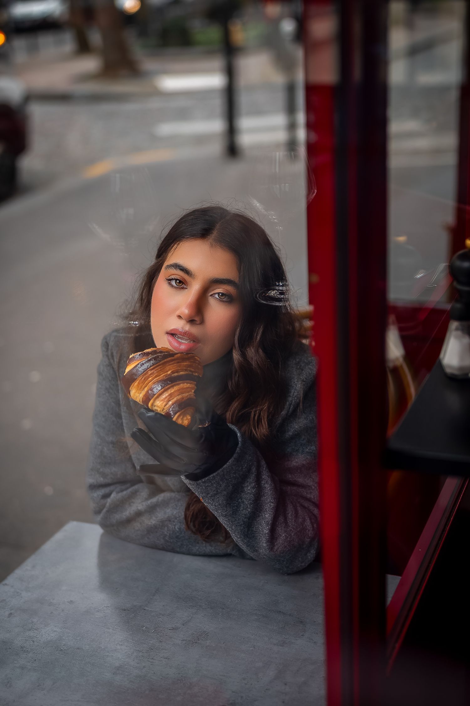 Pensive person in gray sweater gazing through window at cafe table on city street.