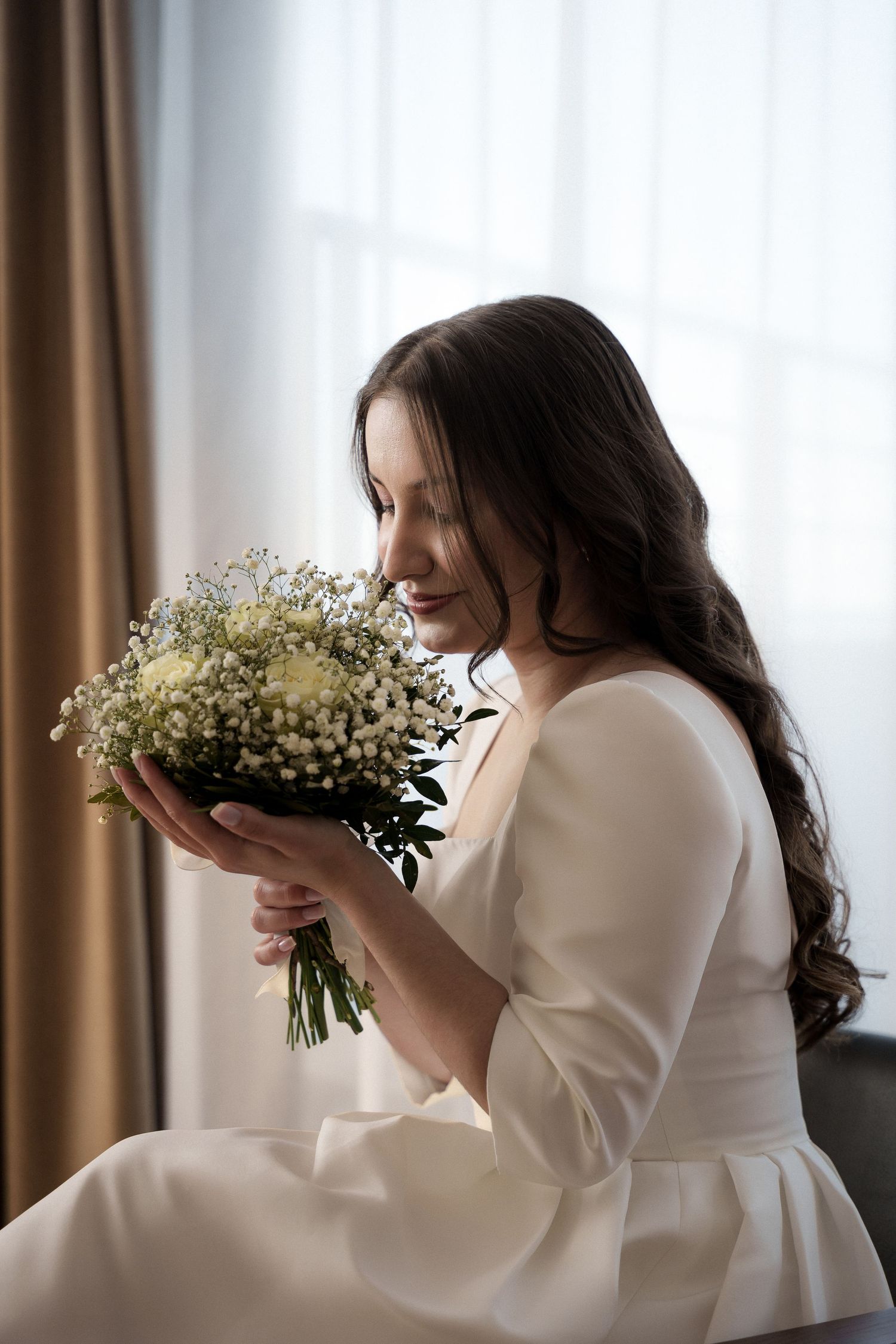 A person in white holds and smells a delicate bouquet of baby's breath flowers near a window with brown curtains.