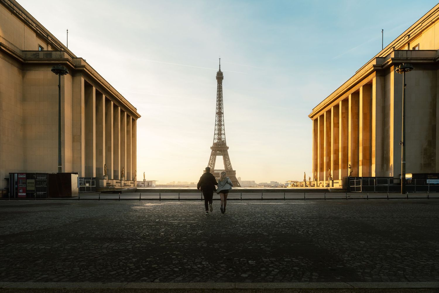Two people walk toward the Eiffel Tower at sunrise, framed by grand colonnades of the Trocadero in Paris.