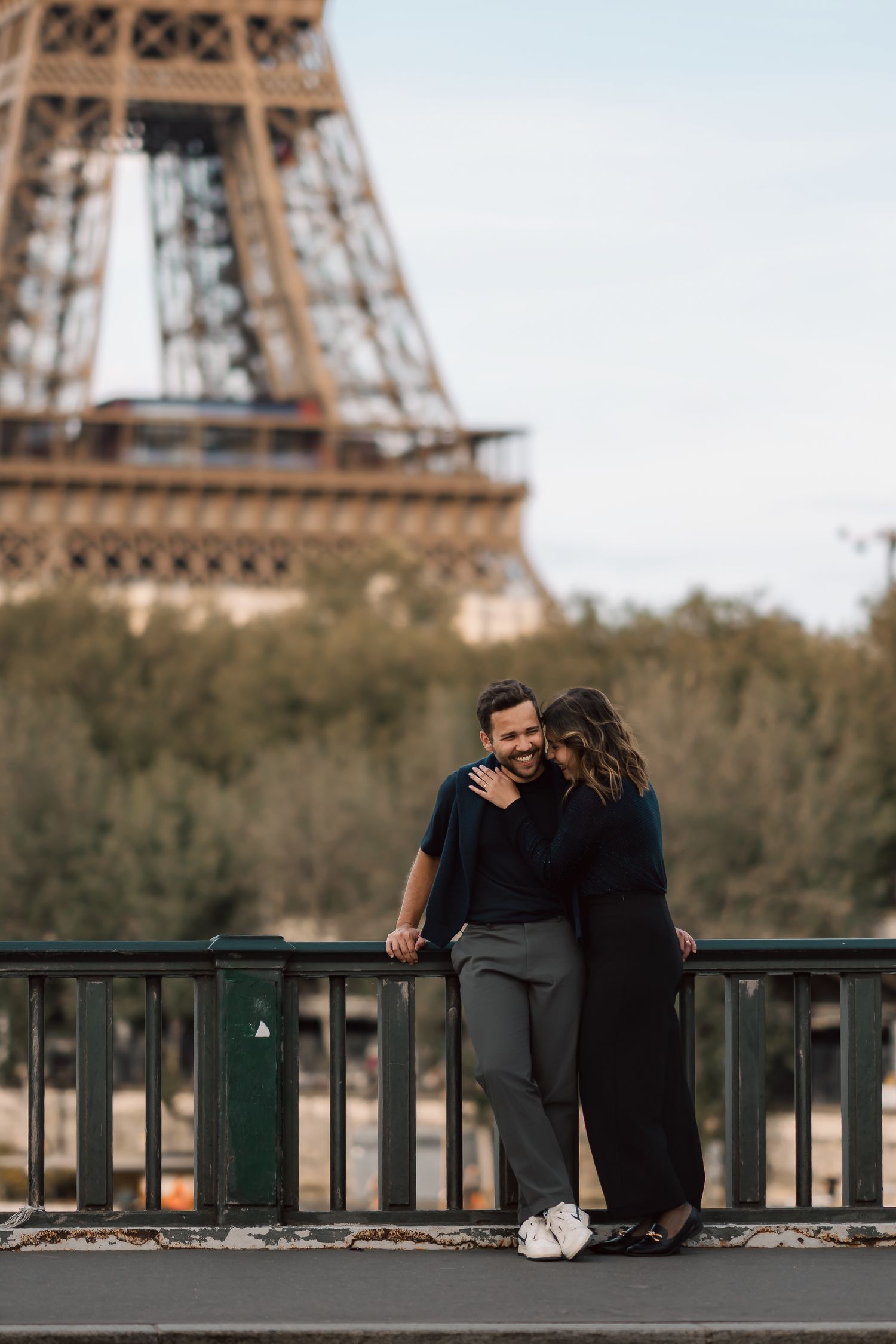 Couple sharing intimate moment on bridge with Eiffel Tower in background.