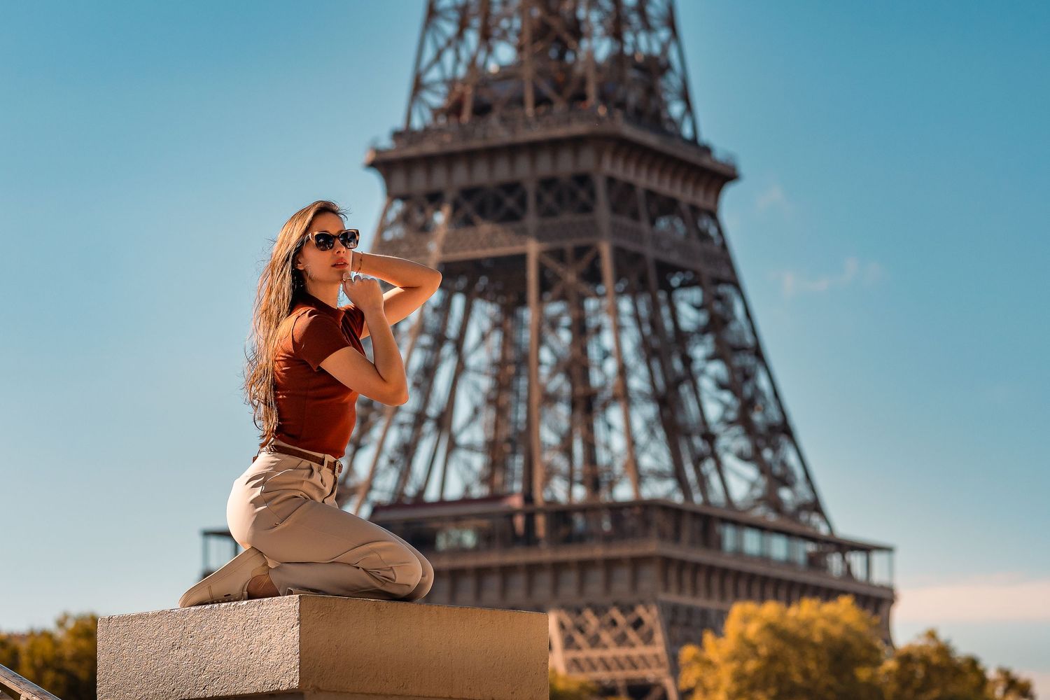 Person sitting on stone ledge poses in front of iconic Eiffel Tower.