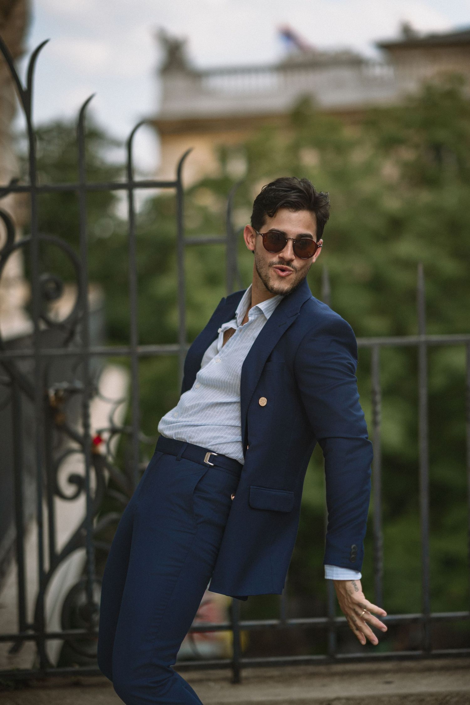 Stylish person in navy suit and sunglasses poses playfully against iron railing in outdoor setting.