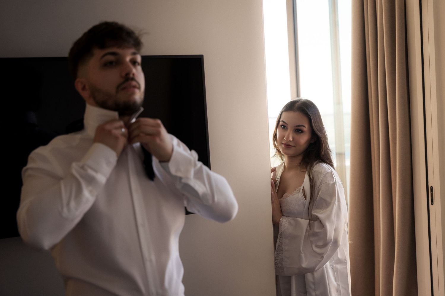 Two people in white shirts stand together in a room with beige curtains and natural lighting.