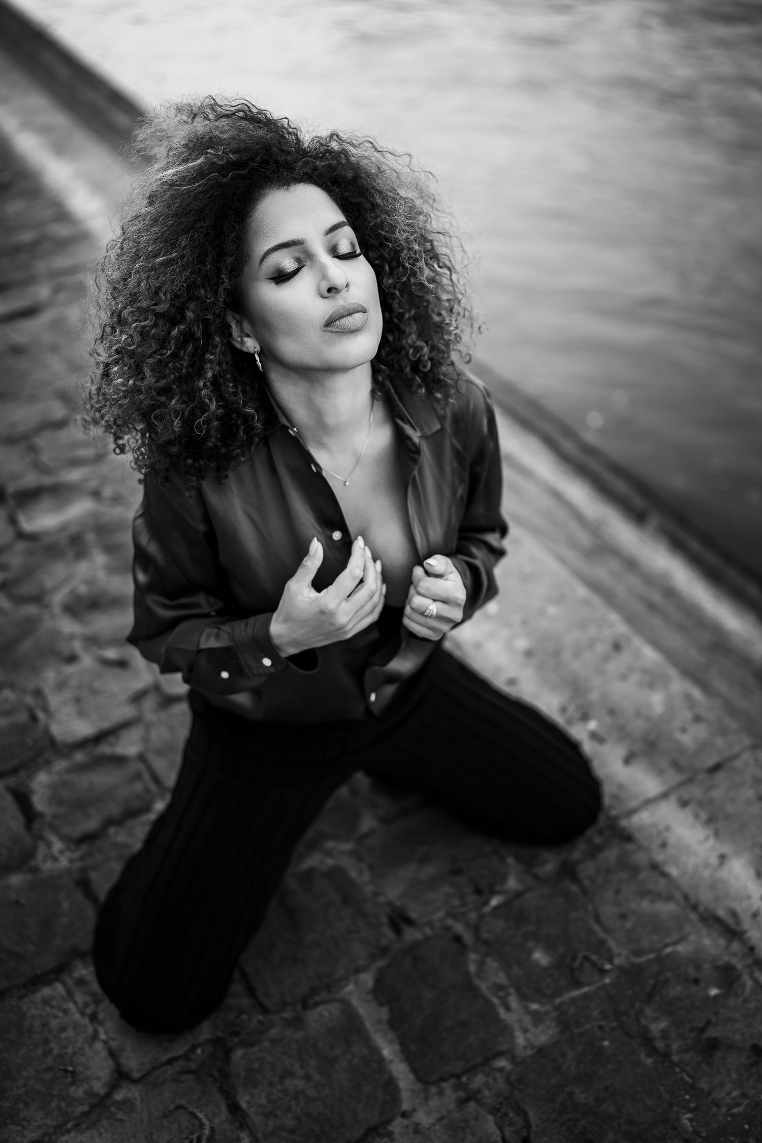 Artistic black and white portrait of someone with curly hair in a dark jacket posing on cobblestone ground.