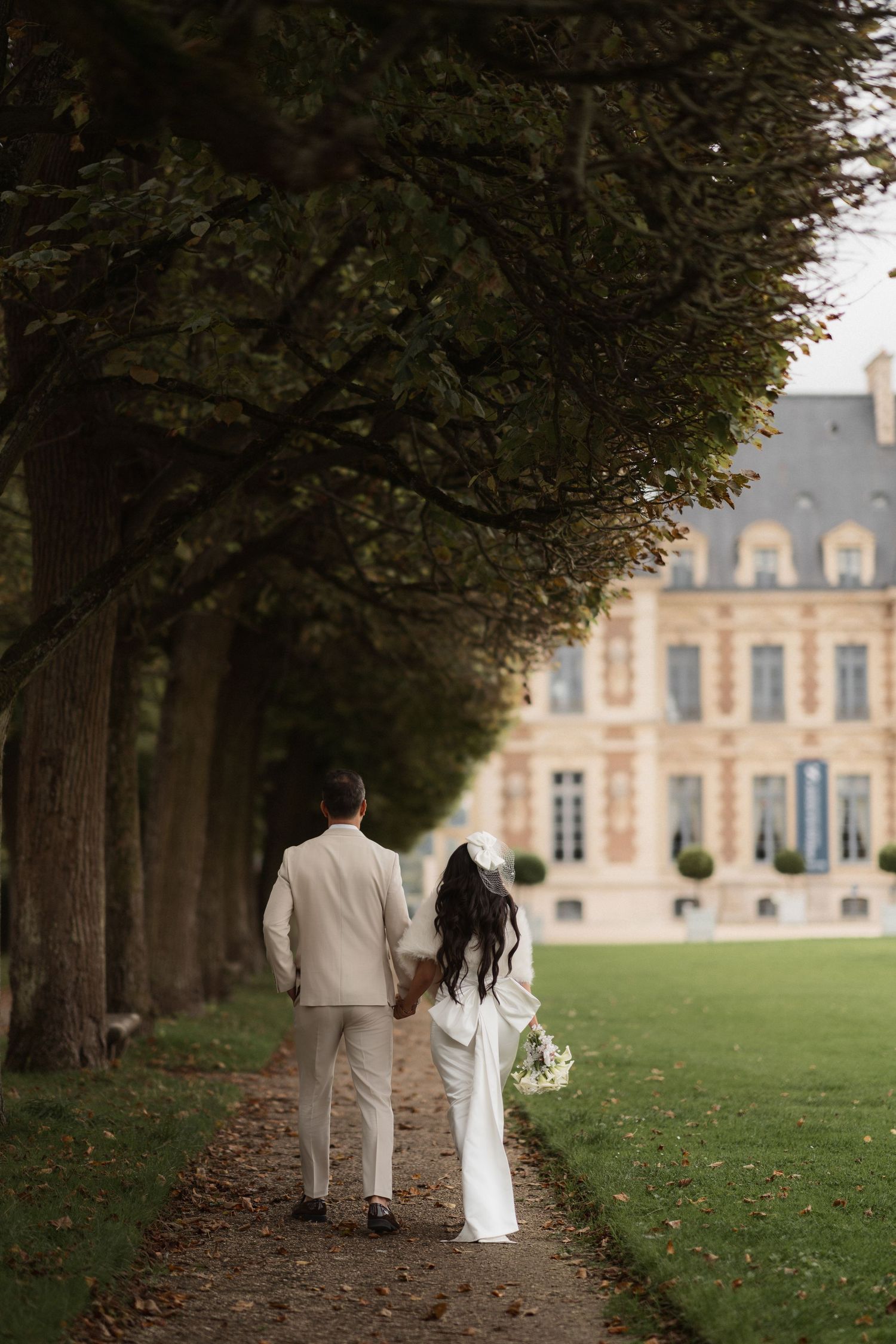A couple in white attire walks down a tree-lined path towards an elegant French chateau on a romantic autumn day.