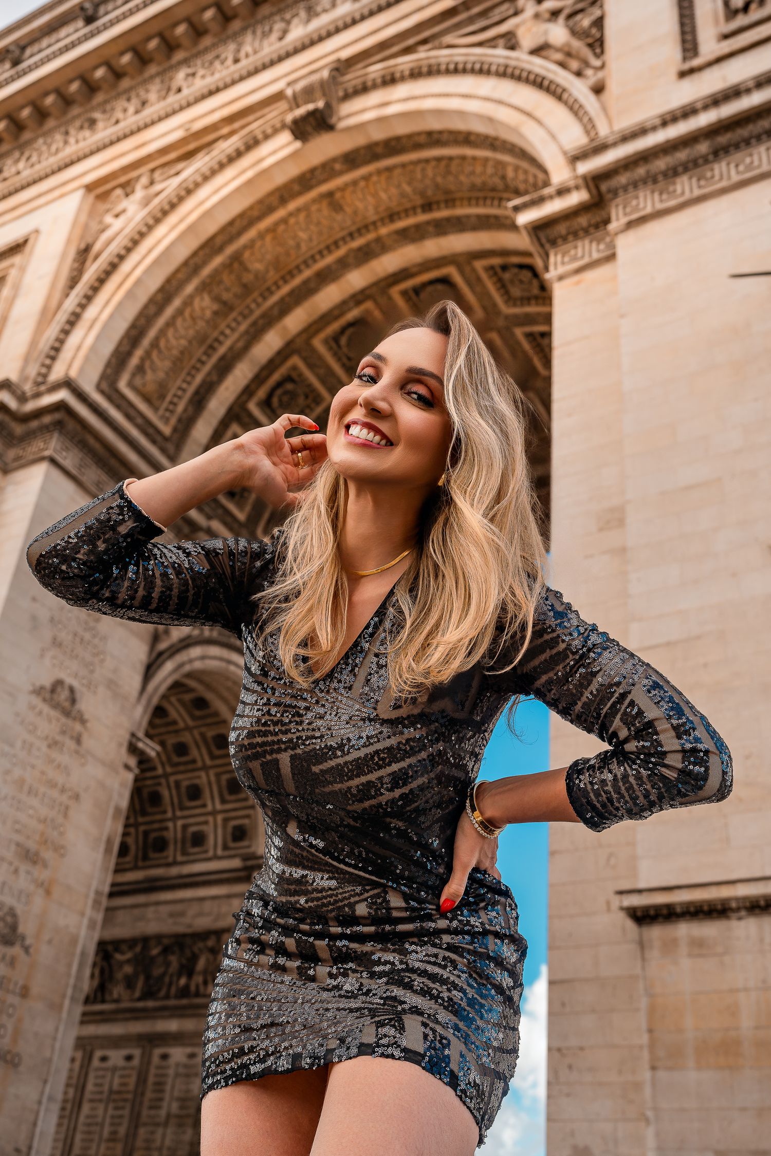Person in a sparkly dress poses joyfully in front of a grand Parisian archway.