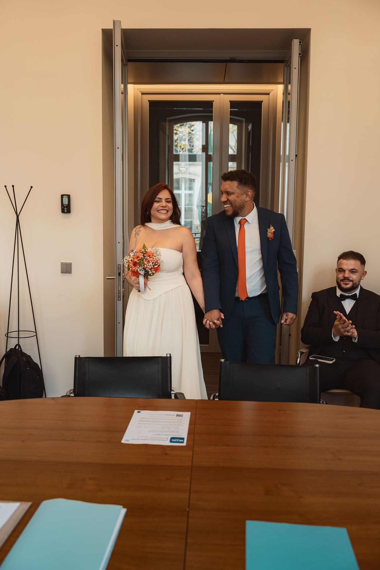 Wedding party members share joyful reactions while gathered around a long wooden conference table in an office setting.