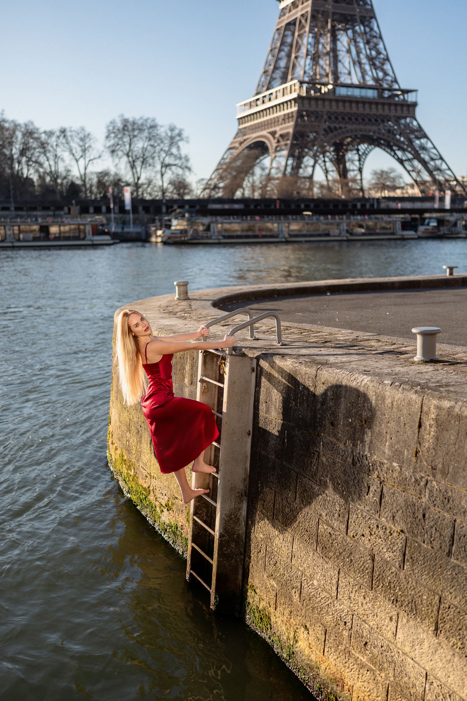 Woman in red dress climbing down ladder by Seine River with Eiffel Tower in background.