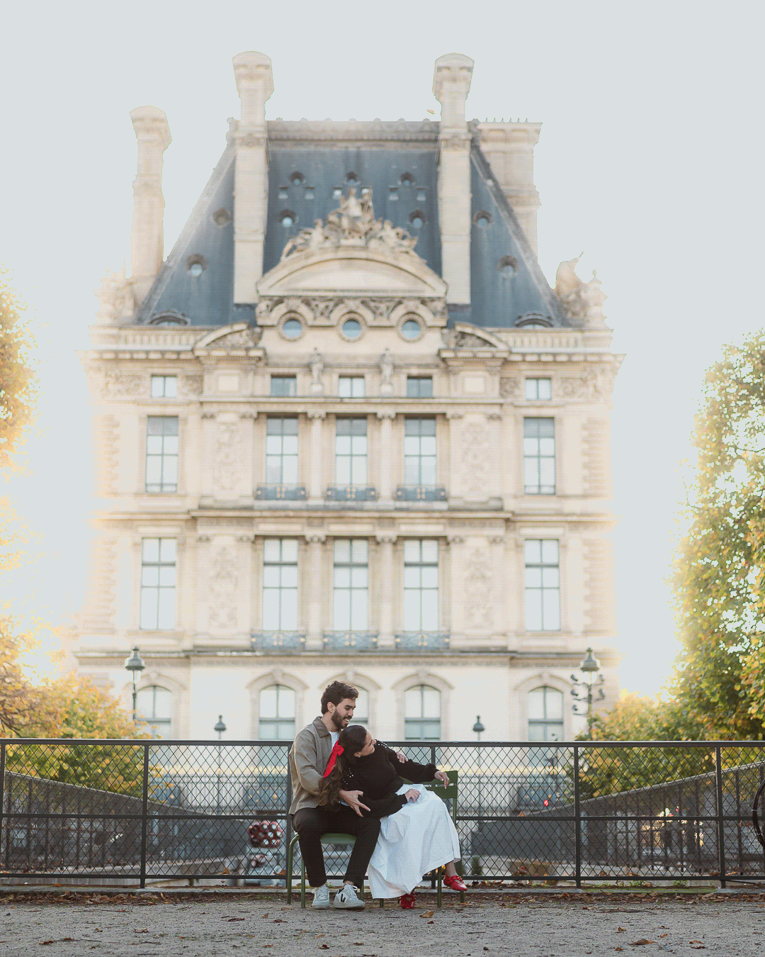 A couple sits together on a bench overlooking the grand facade of the Louvre Museum in Paris at sunset.