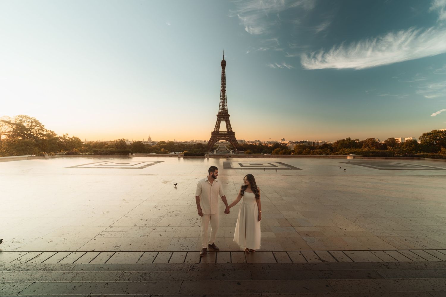 A romantic couple walks across the Trocadero plaza with the iconic Eiffel Tower in the background at sunset.