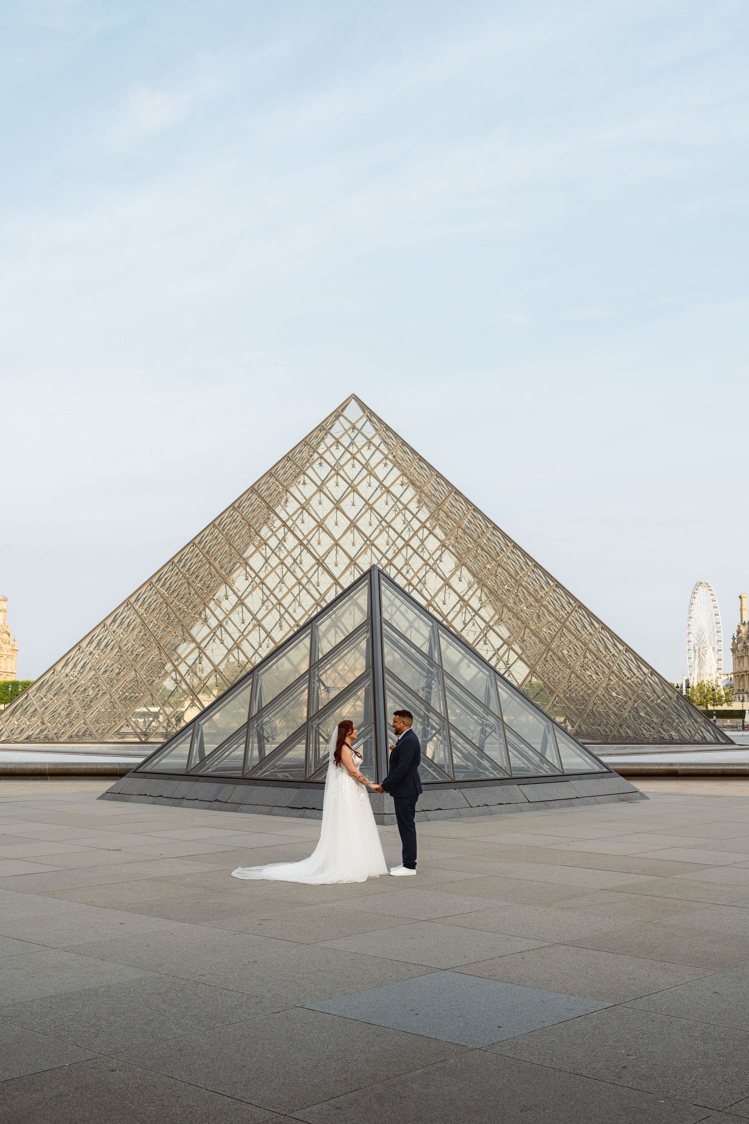 A couple poses for wedding photos in front of the iconic glass pyramid at the Louvre Museum in Paris at sunset.