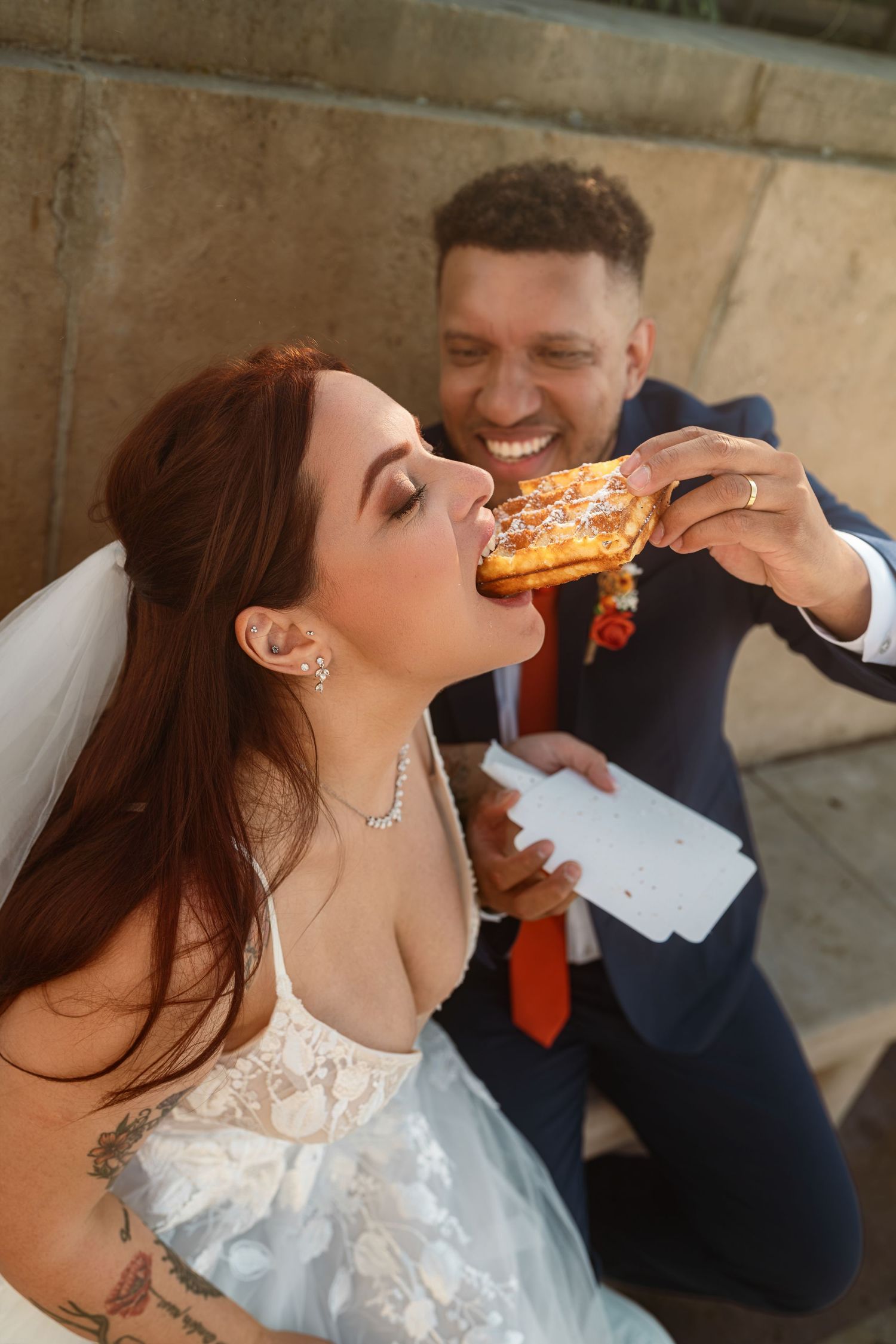 A couple shares pizza together on their wedding day, with the bride taking playful bites in her white lace dress.