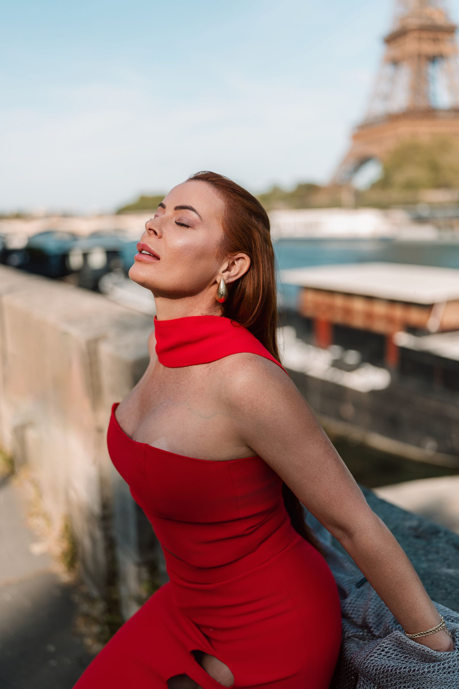 Person in red dress poses against Seine River railing with Eiffel Tower in background.