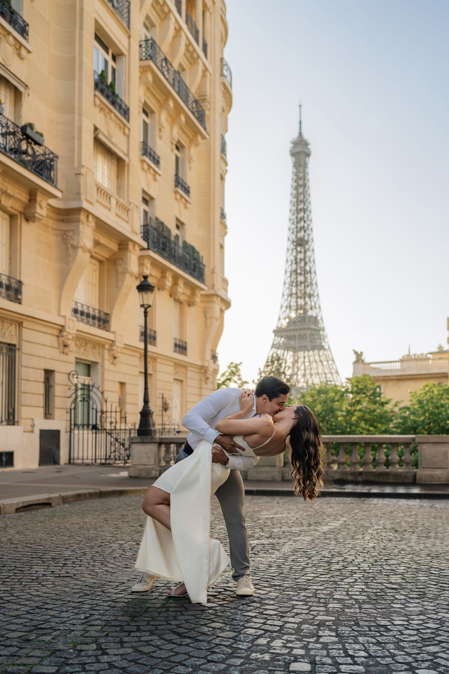 Couple in elegant attire sharing romantic dip kiss on Paris street with Eiffel Tower view.