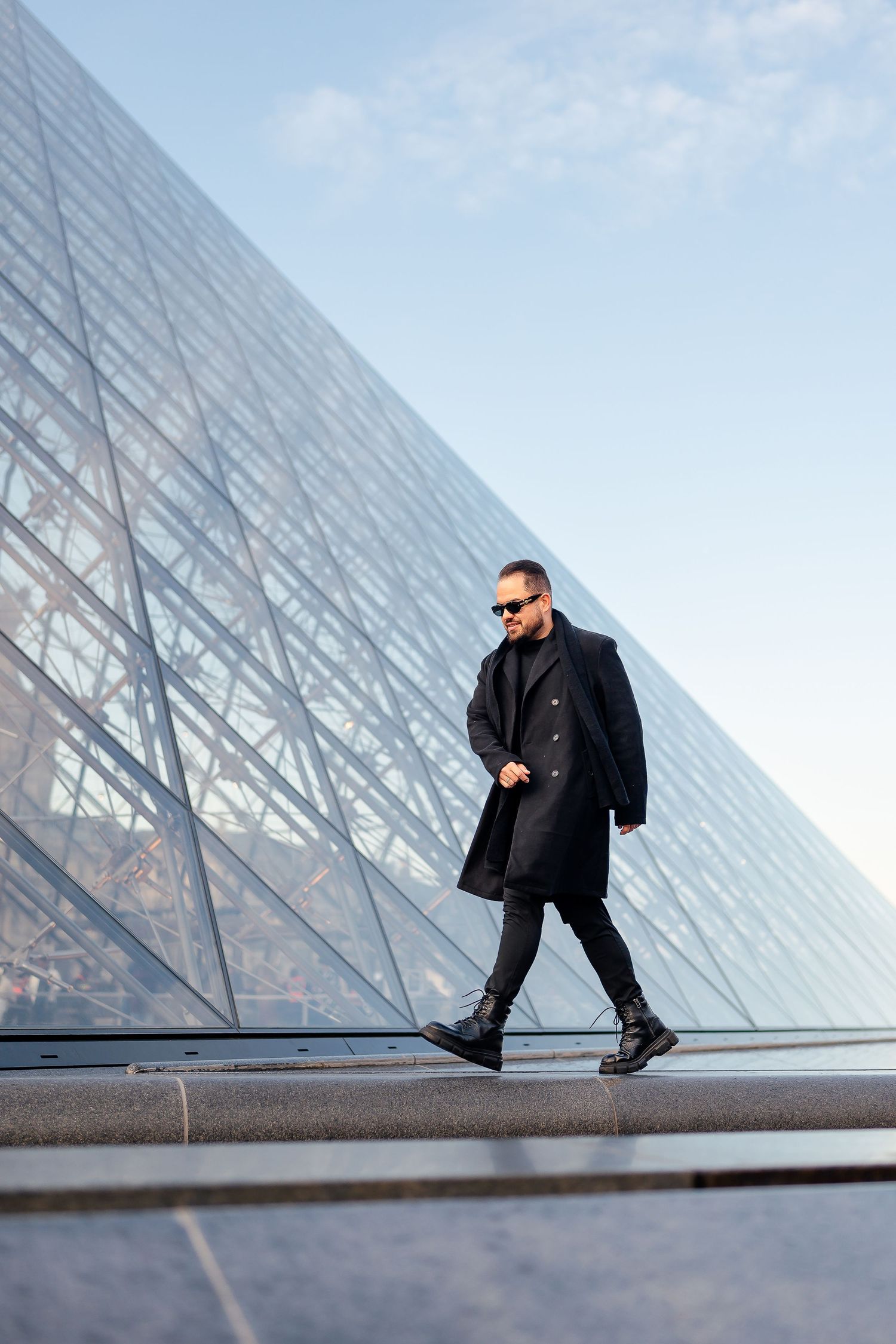Person in black clothing and sunglasses walks along a geometric glass structure in an artistic architectural setting.