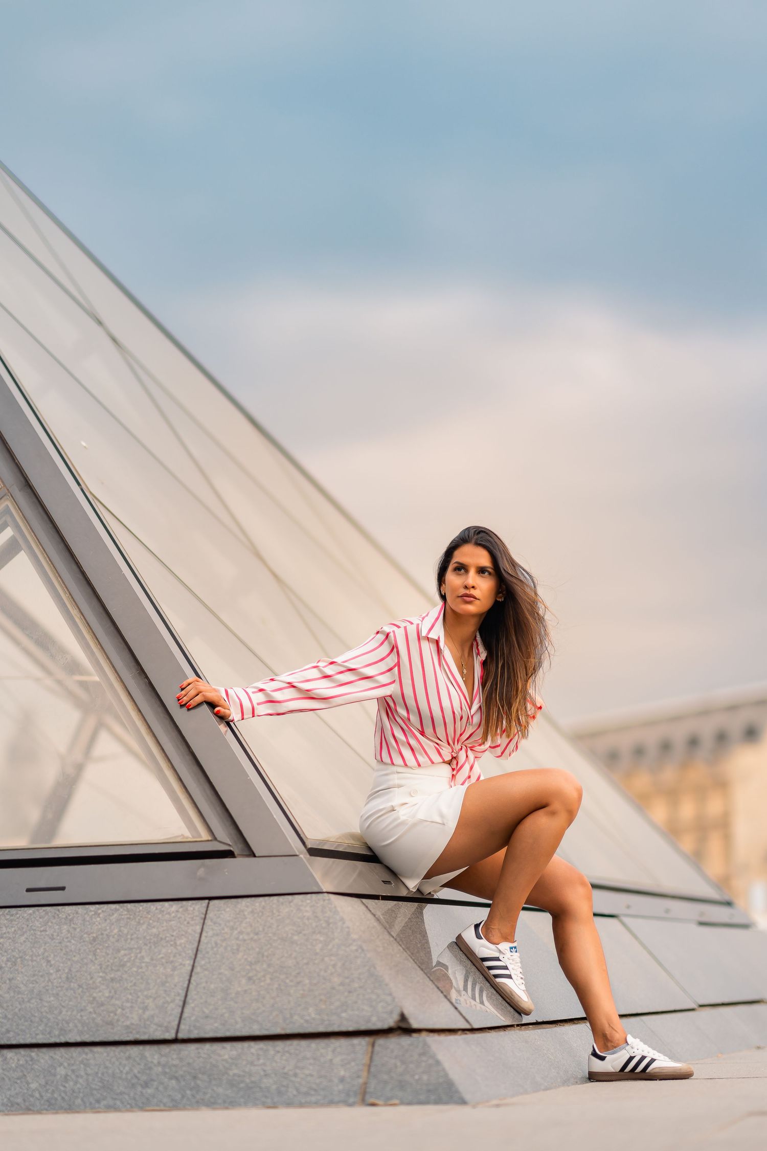 A person in a striped top and white shorts poses on an angled architectural surface against a cloudy sky.