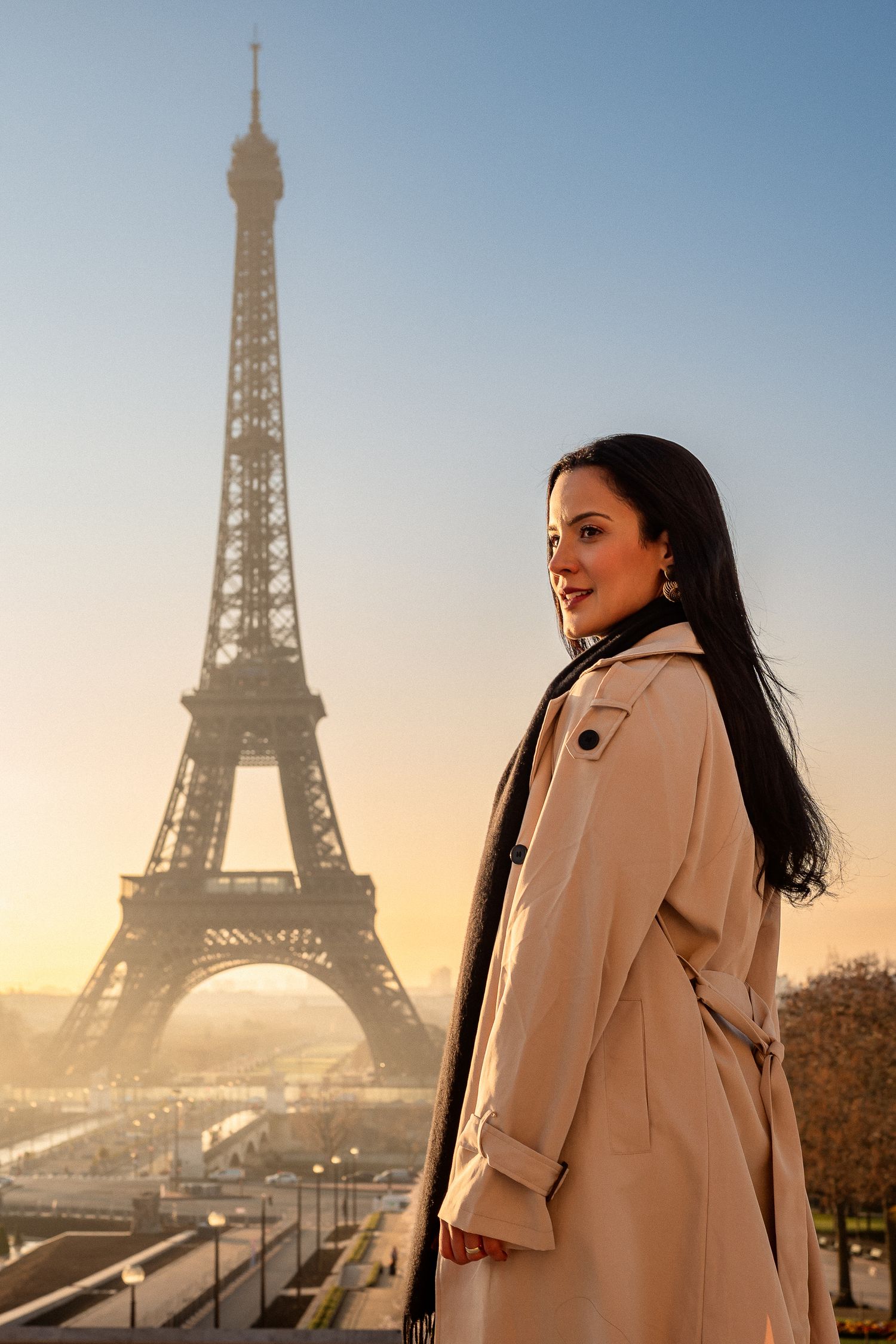 Person in beige coat poses against sunset sky with Eiffel Tower silhouette.
