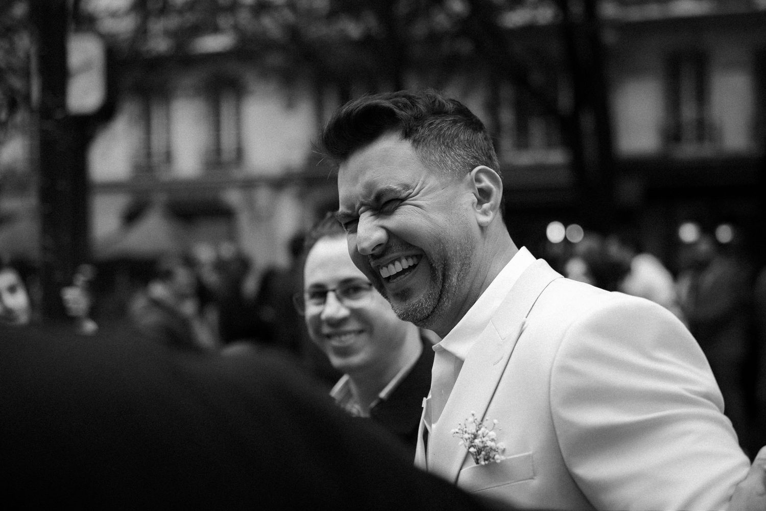 A candid black and white photo showing joyful laughter at an outdoor event, with one person wearing a white blazer.