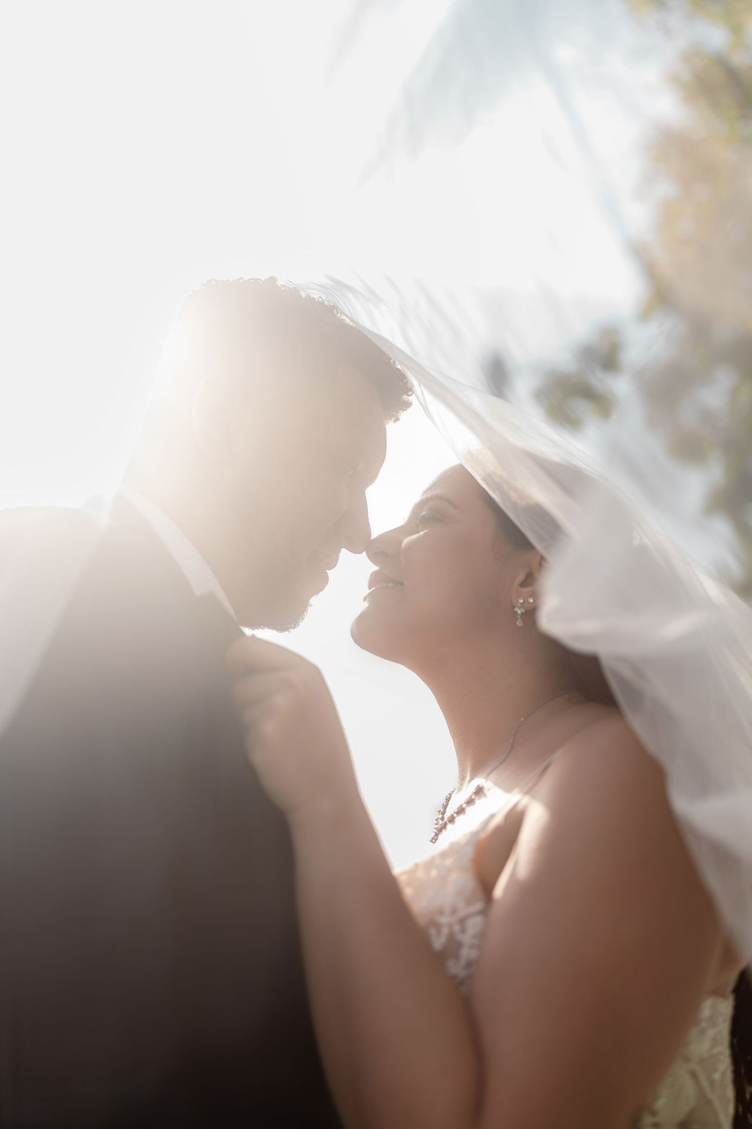 Romantic silhouette of a couple under a delicate white veil with soft sunlight filtering through in an ethereal moment.