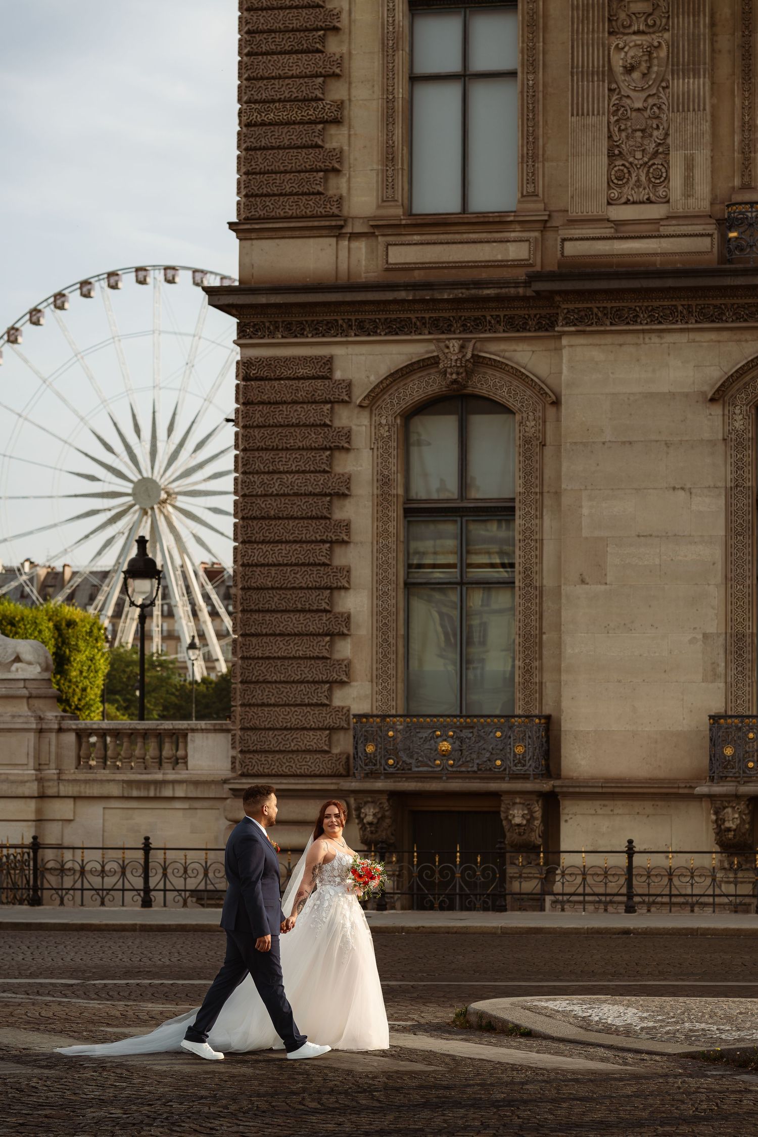 A couple in formal wedding attire walks along a historic building facade with a Ferris wheel visible in the background.