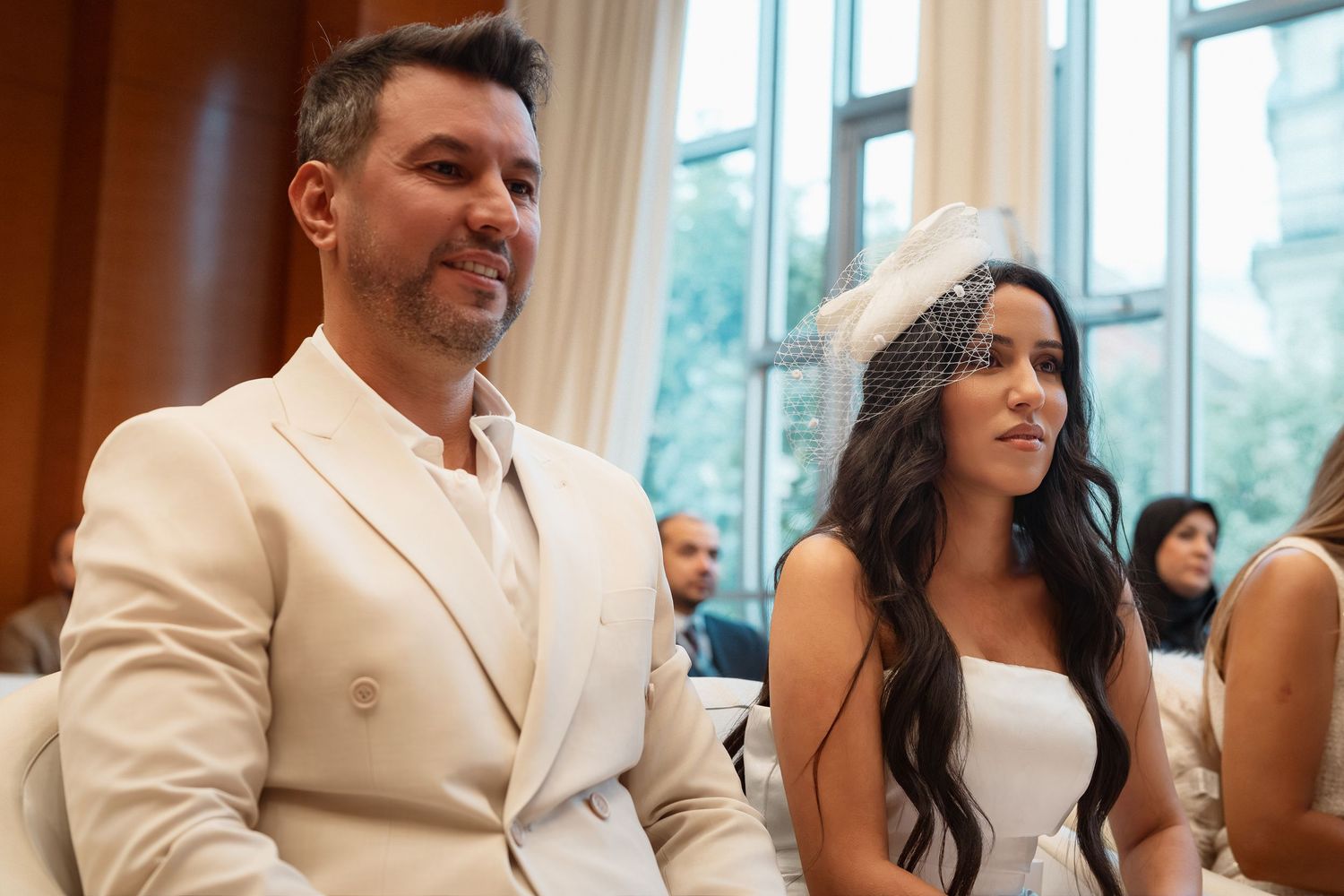 Wedding guests in cream-colored attire smile and laugh during an elegant indoor celebration.