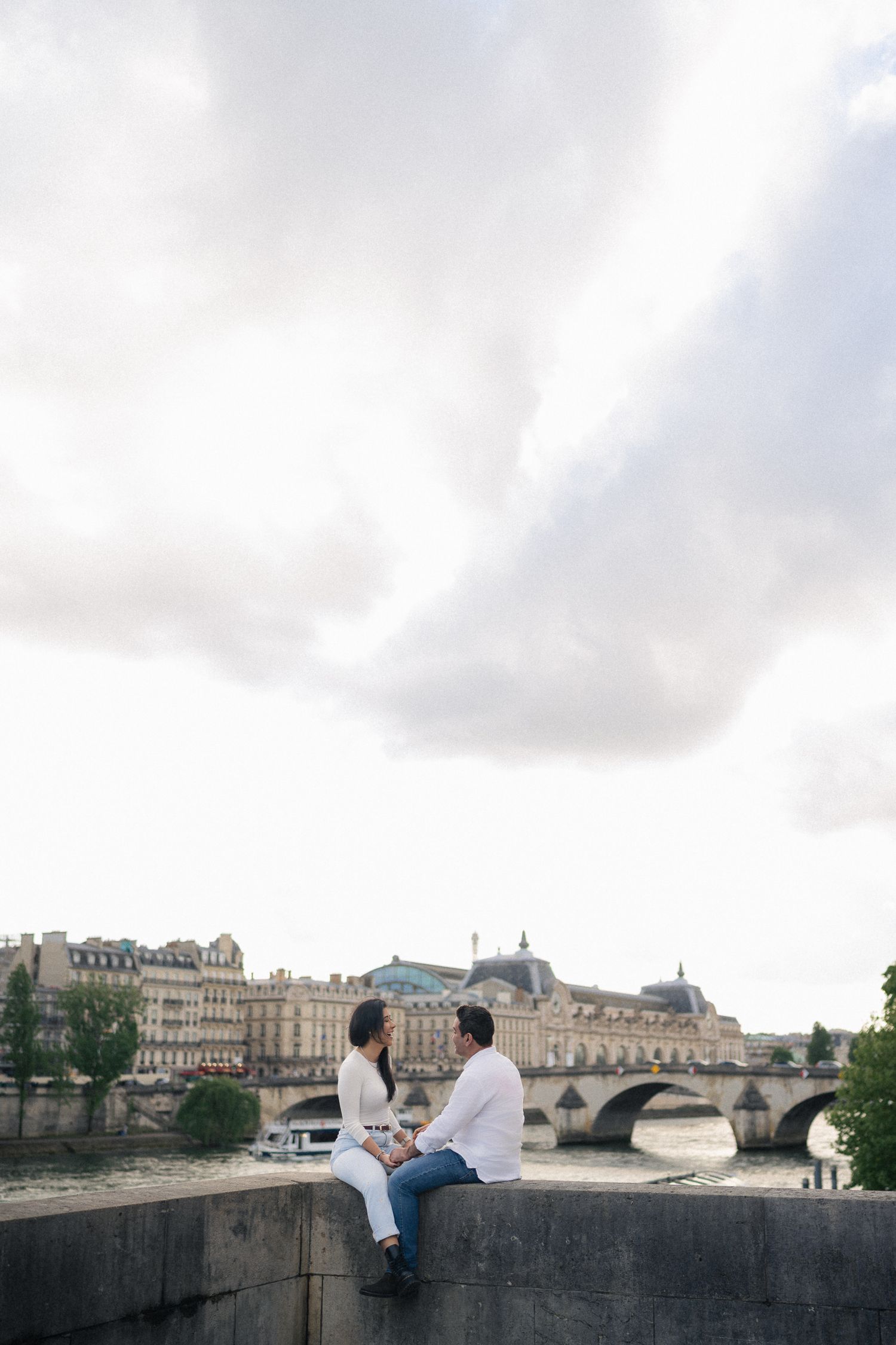 A couple sits on a stone wall overlooking the Seine River and Pont Neuf bridge in Paris.