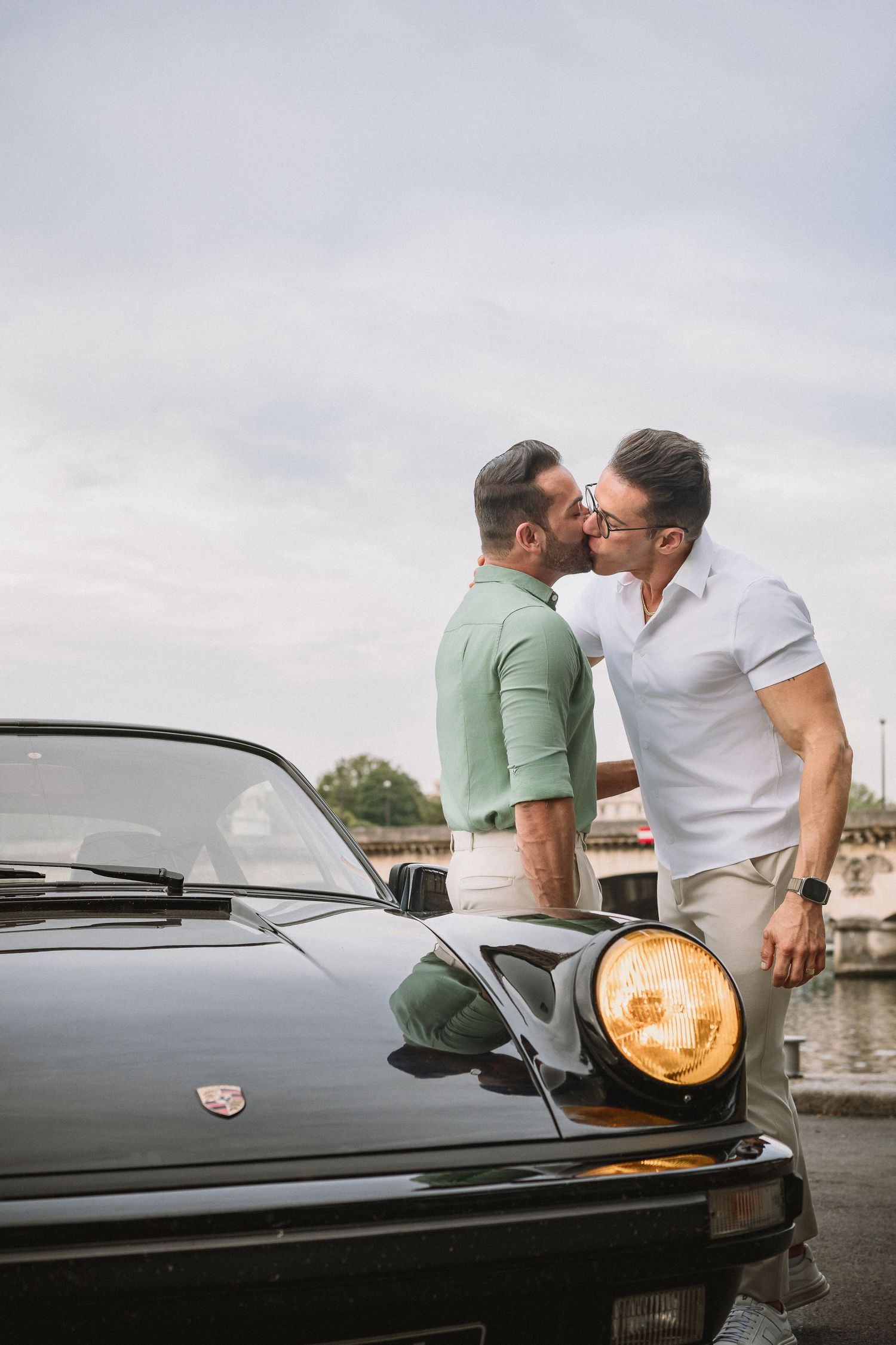 A couple shares an intimate moment while sitting on a vintage black car by the waterfront at dusk.